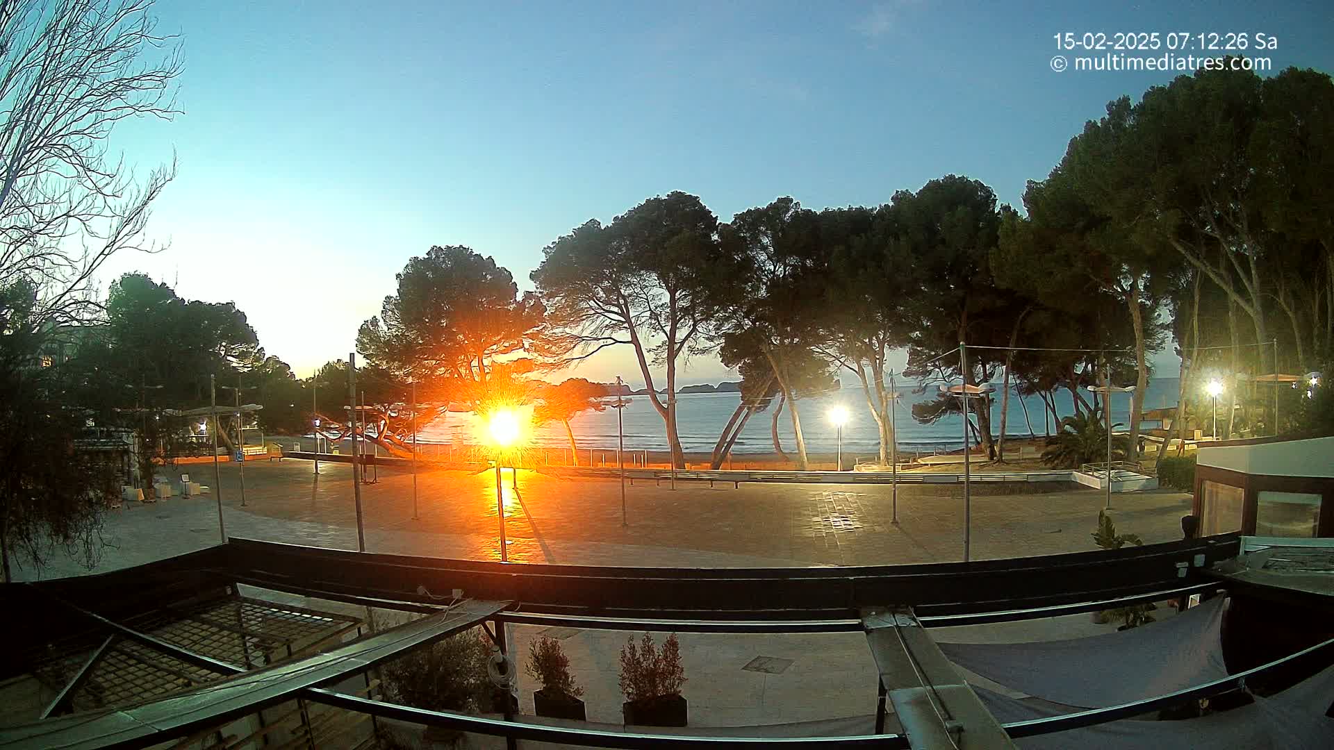 A sunrise over a calm ocean, viewed from a paved plaza lined with trees and lit by streetlights.
