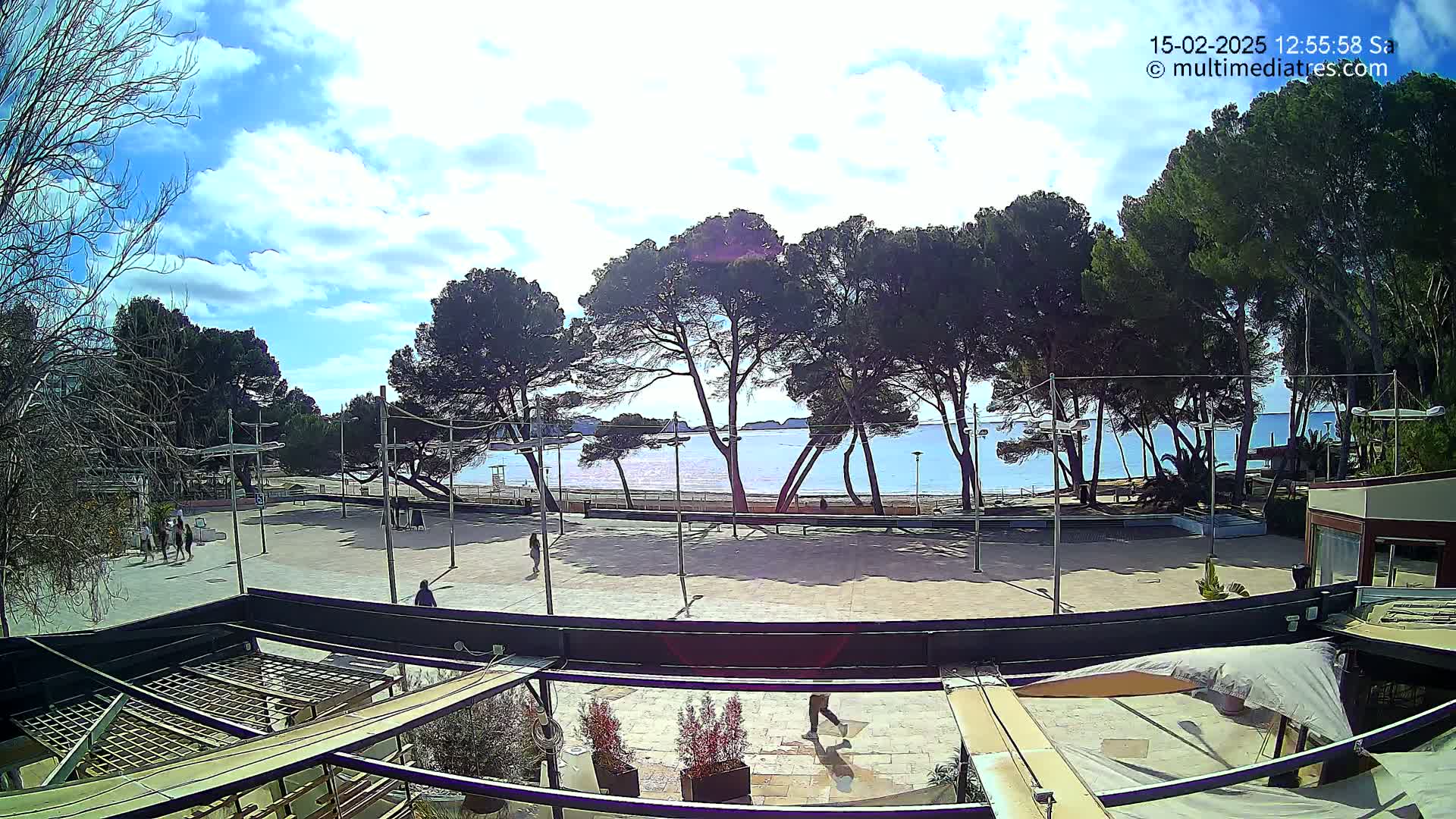 A partly sunny day reveals a seaside plaza with several people walking, trees lining a sandy beach, and calm blue water in the distance.