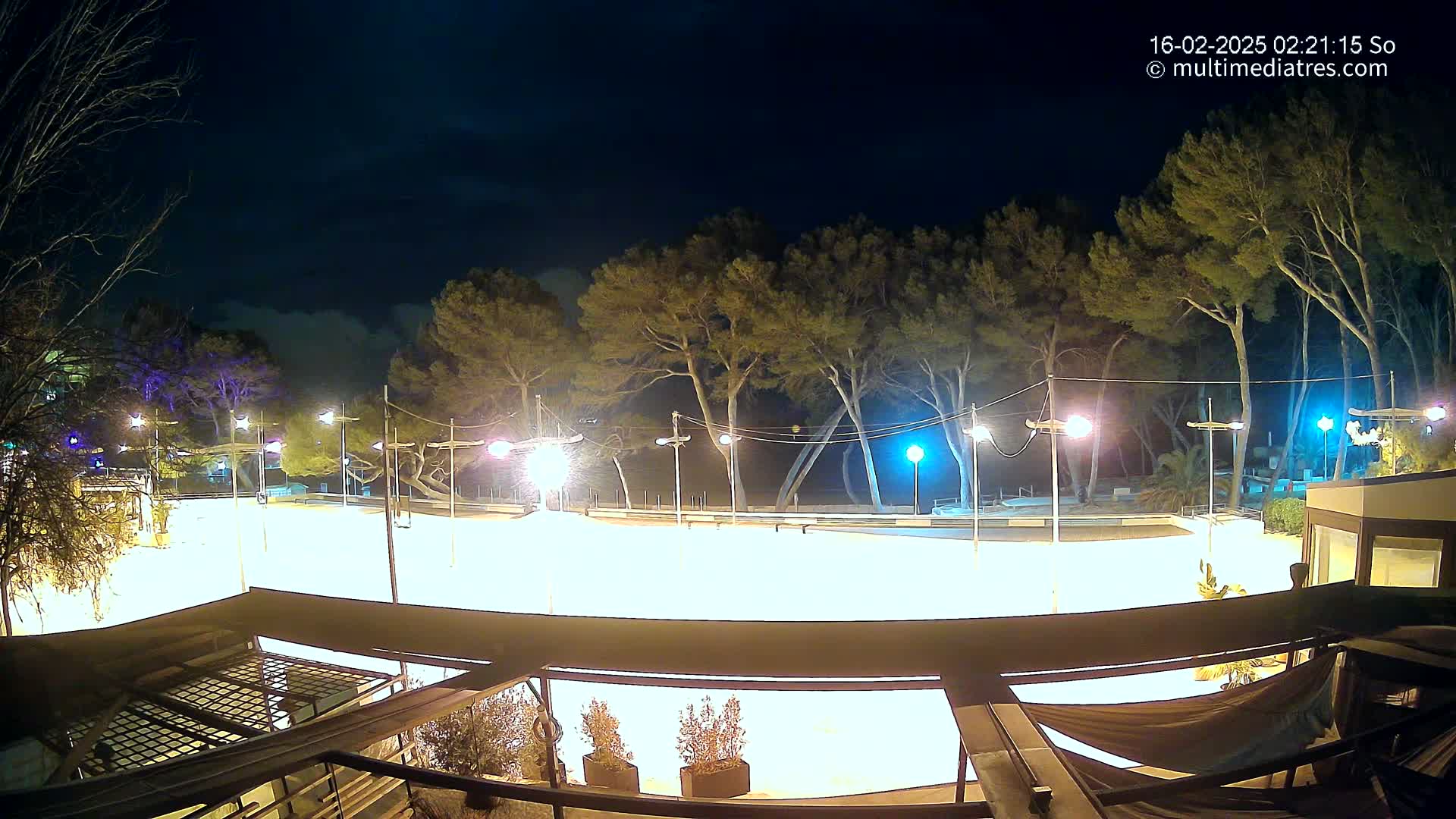 A brightly lit, empty outdoor rink is surrounded by trees under a partly cloudy night sky.