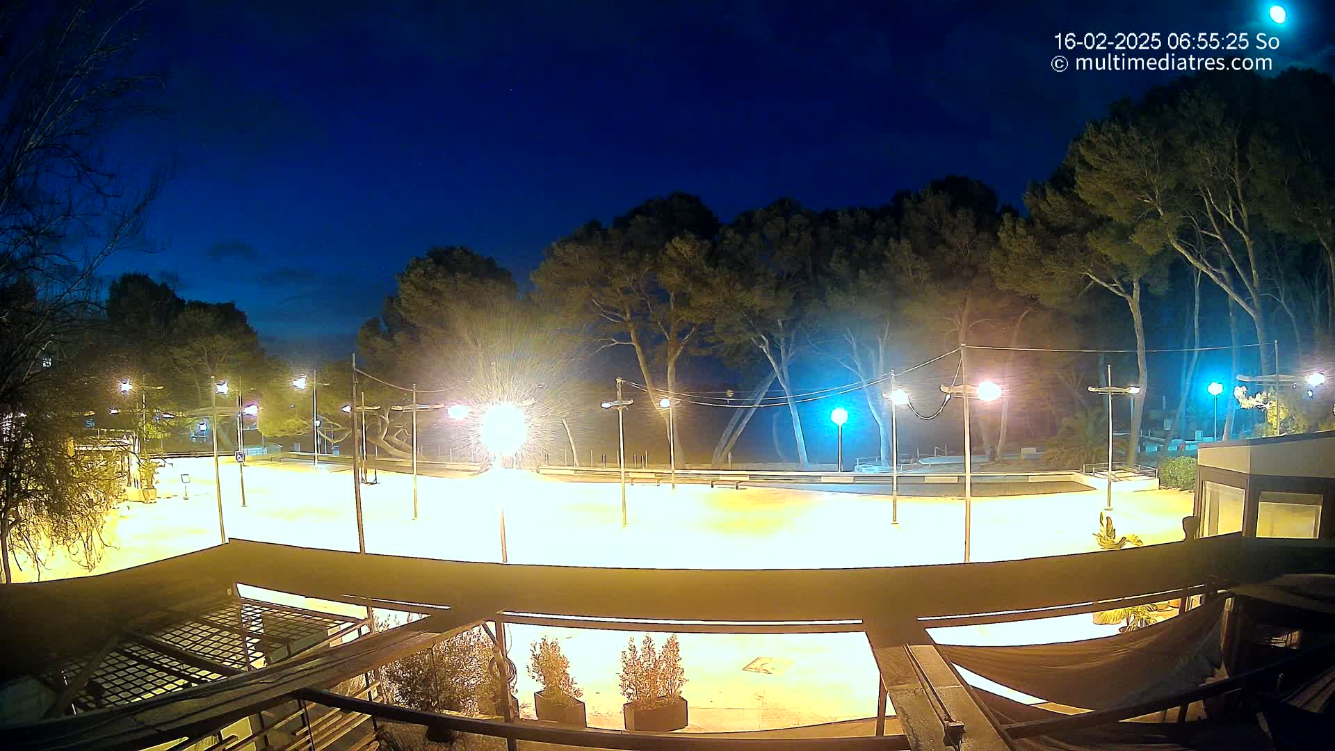 A brightly lit, empty plaza at night, bordered by trees under a clear, dark blue sky.