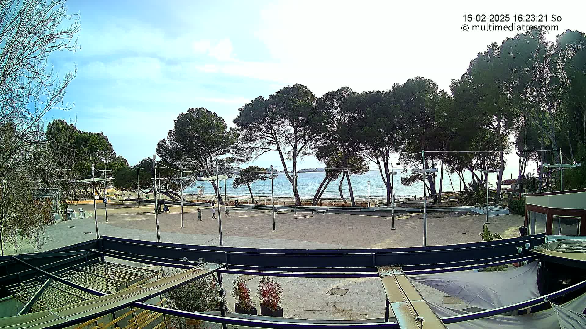 A mostly sunny day overlooking a paved plaza next to a sandy beach with calm water and a line of pine trees.