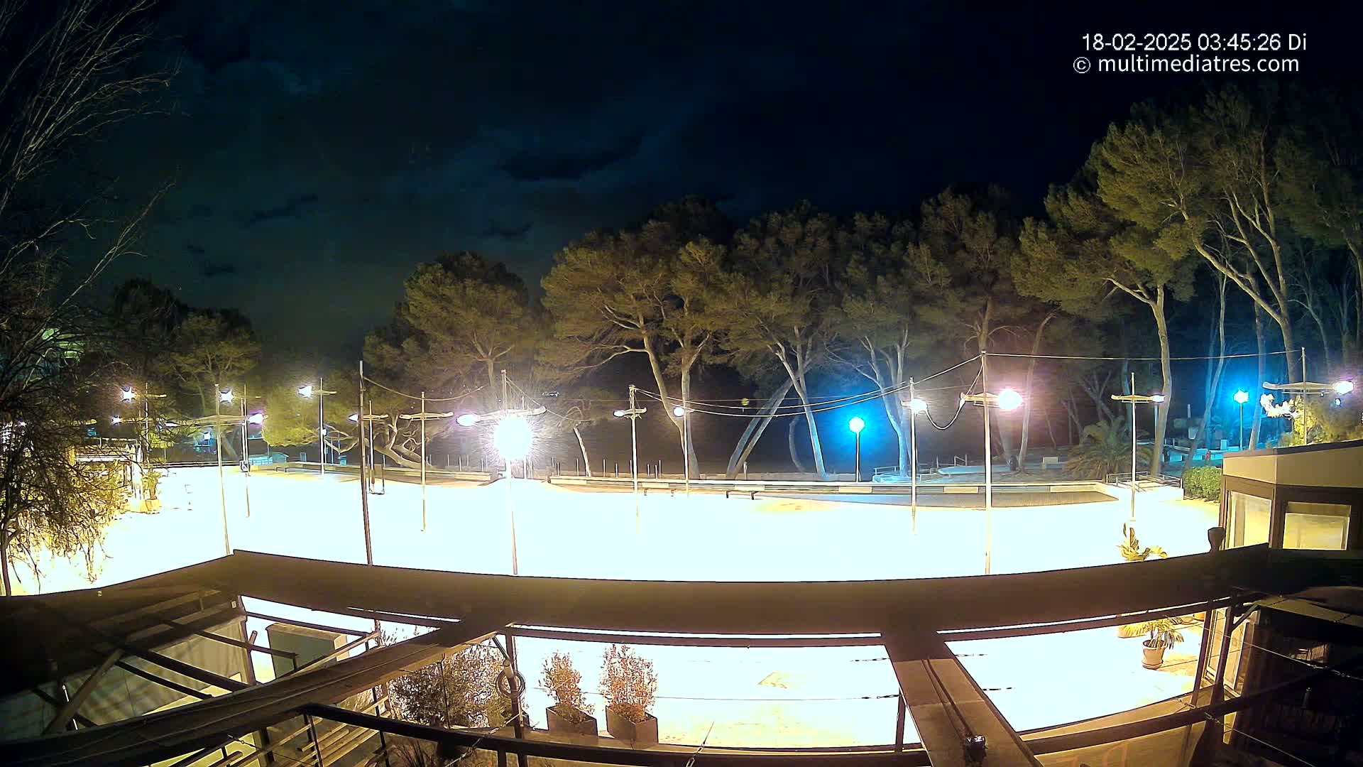 A brightly lit, empty paved area at night, surrounded by trees under a partly cloudy sky.