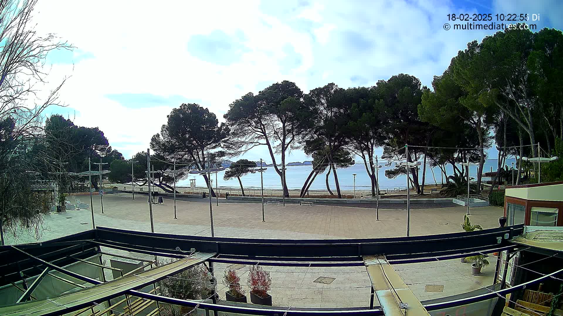 A mostly sunny outdoor view of a paved plaza with trees, leading to a calm beach and ocean under a partly cloudy sky.