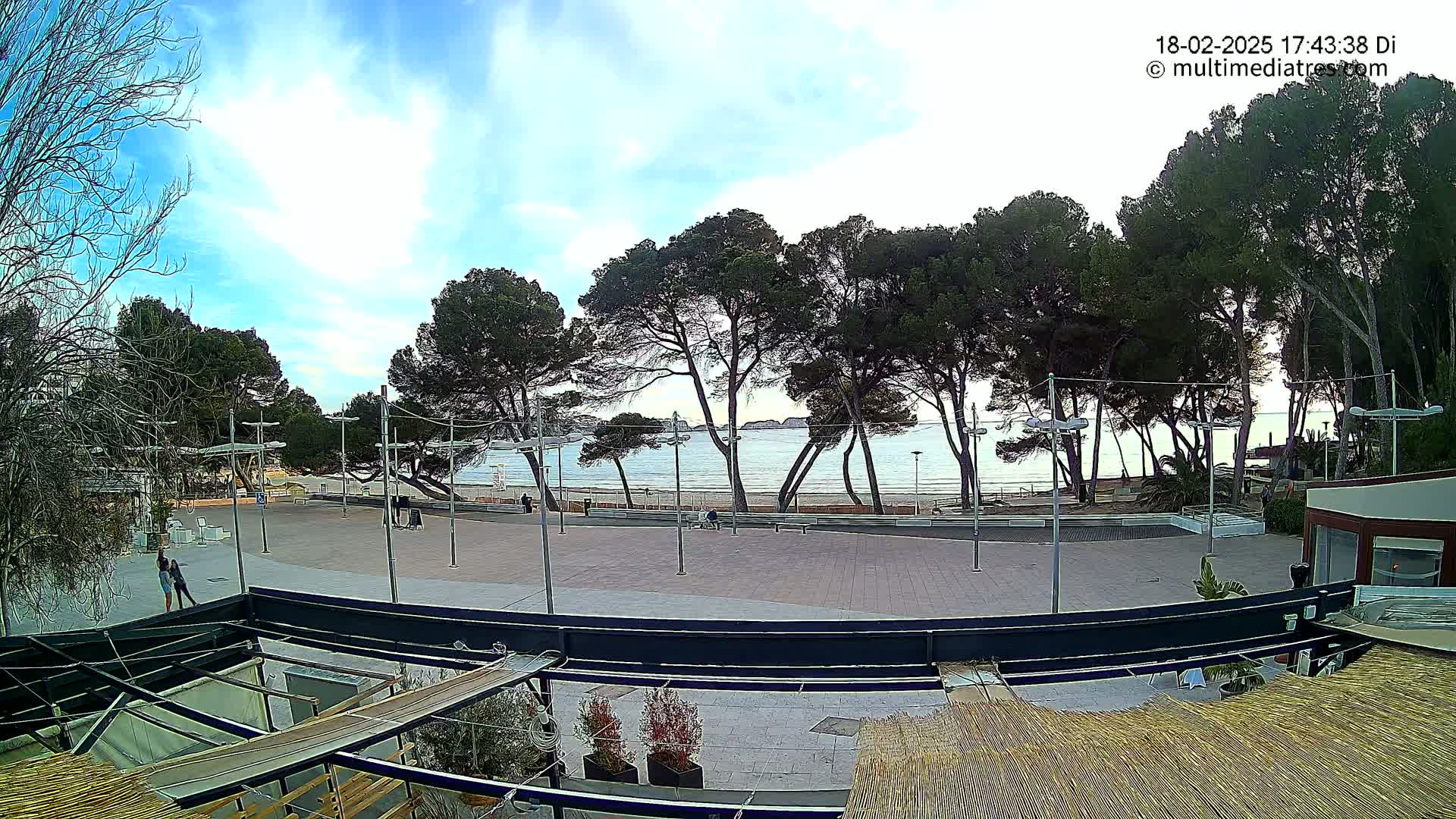 A paved plaza overlooks a calm sea with a sandy beach and a line of pine trees under a partly cloudy sky.