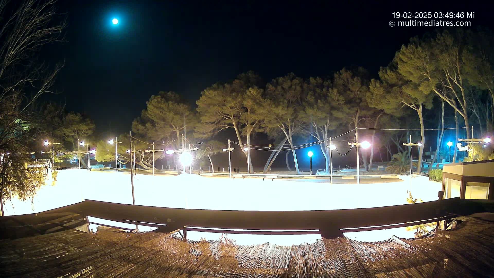 A brightly lit, empty plaza at night under a clear sky with a visible moon, surrounded by tall, dark trees and a building with a thatched roof.