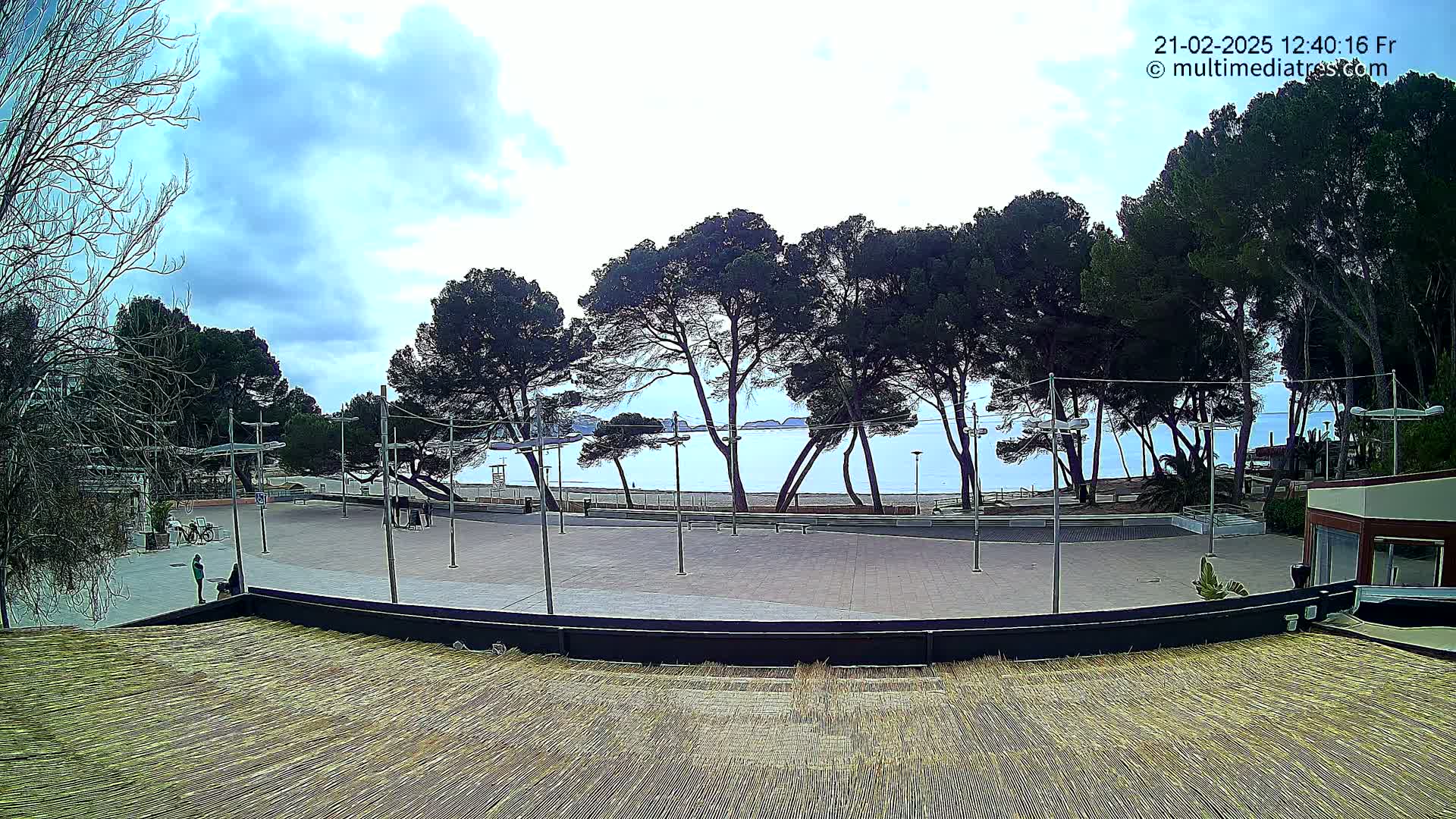 A mostly empty plaza overlooks a calm ocean beach under a partly cloudy sky, with a line of dark green trees separating the two.