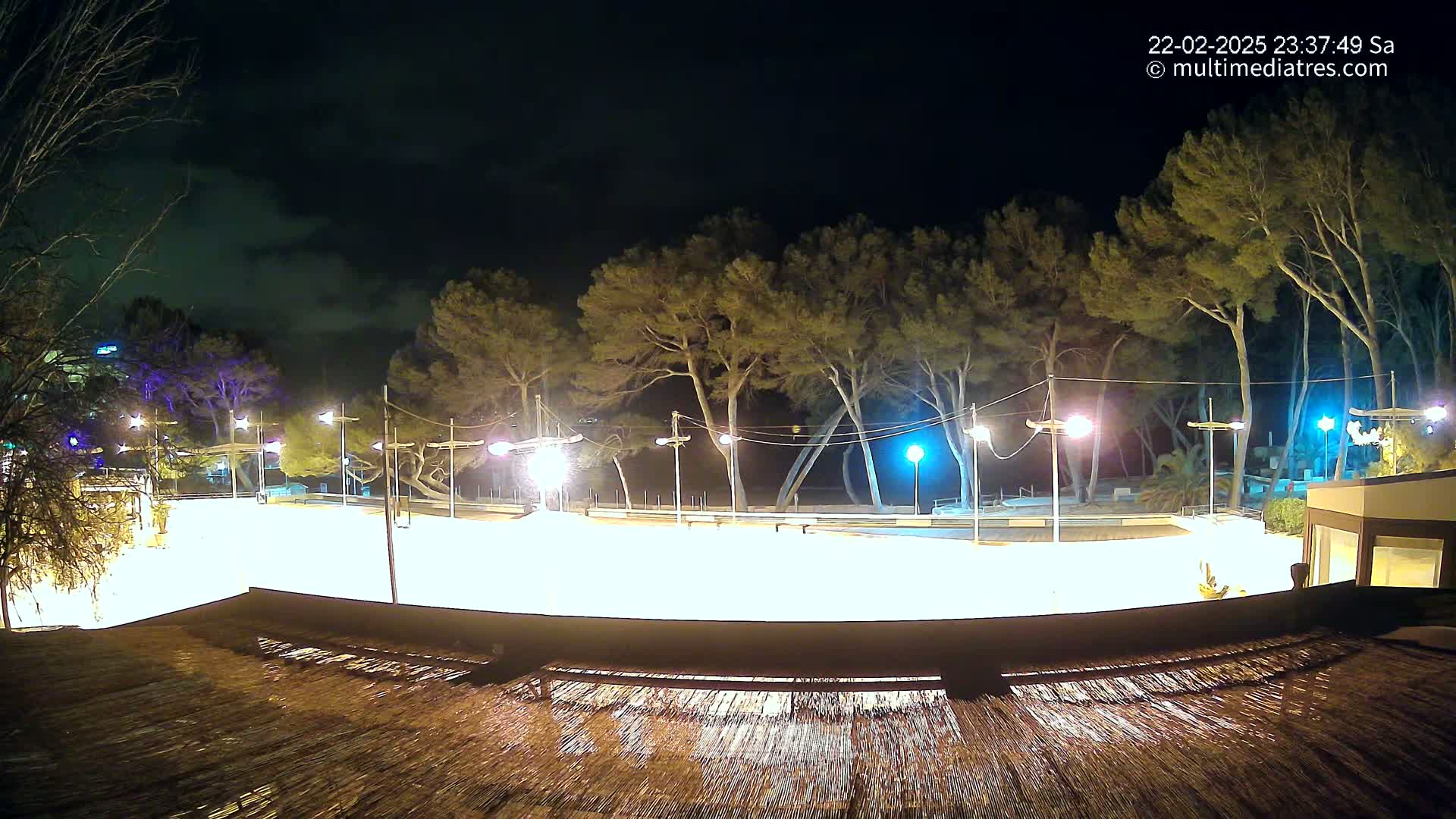 A brightly lit, empty skating rink at night, surrounded by trees under a dark, cloudy sky.