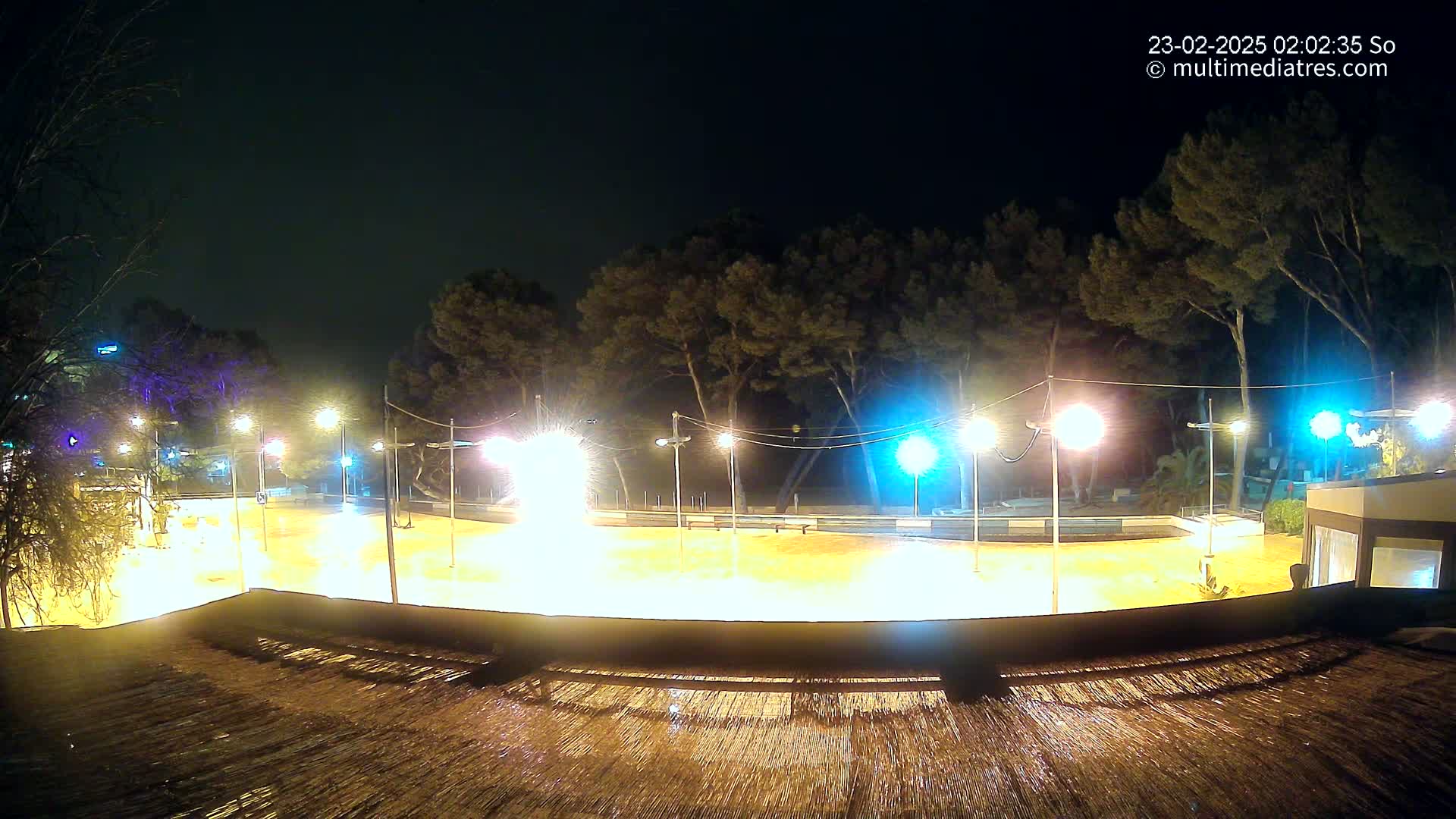 A brightly lit, empty plaza at night, surrounded by trees, is seen from an elevated vantage point, with rain reflecting light on a surface in the foreground.