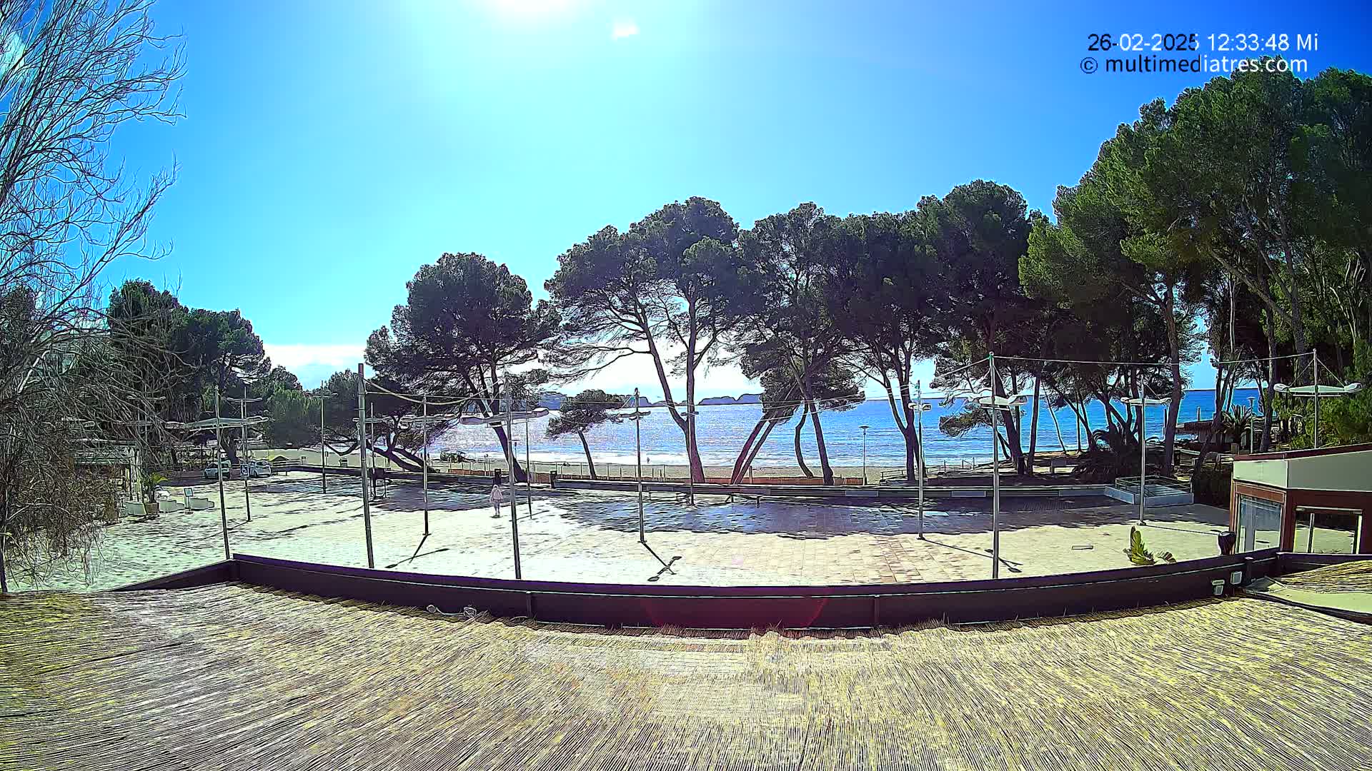 A paved outdoor area, possibly a plaza or court, overlooks a sandy beach and calm ocean under a bright, sunny sky.