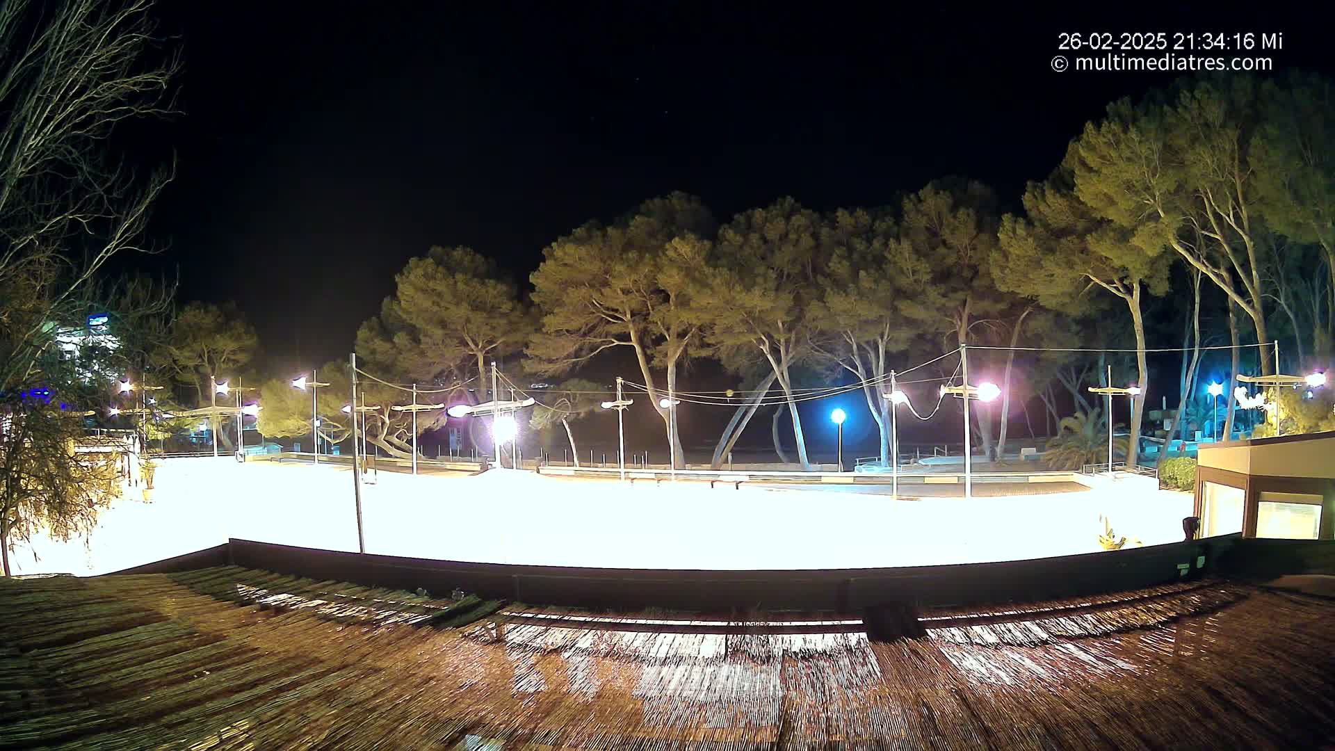 A brightly lit, empty paved area at night, bordered by trees and buildings under a clear, dark sky.