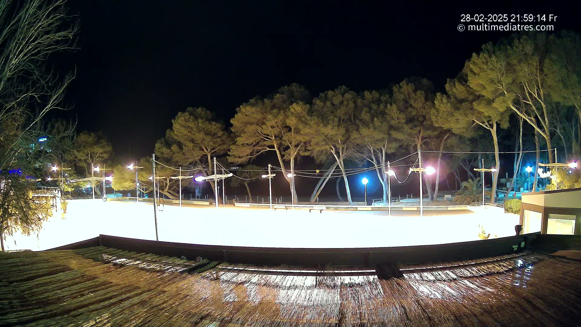 A brightly lit, empty skating rink at night is surrounded by trees and illuminated by numerous streetlights, with a wet, thatched roof visible in the foreground.