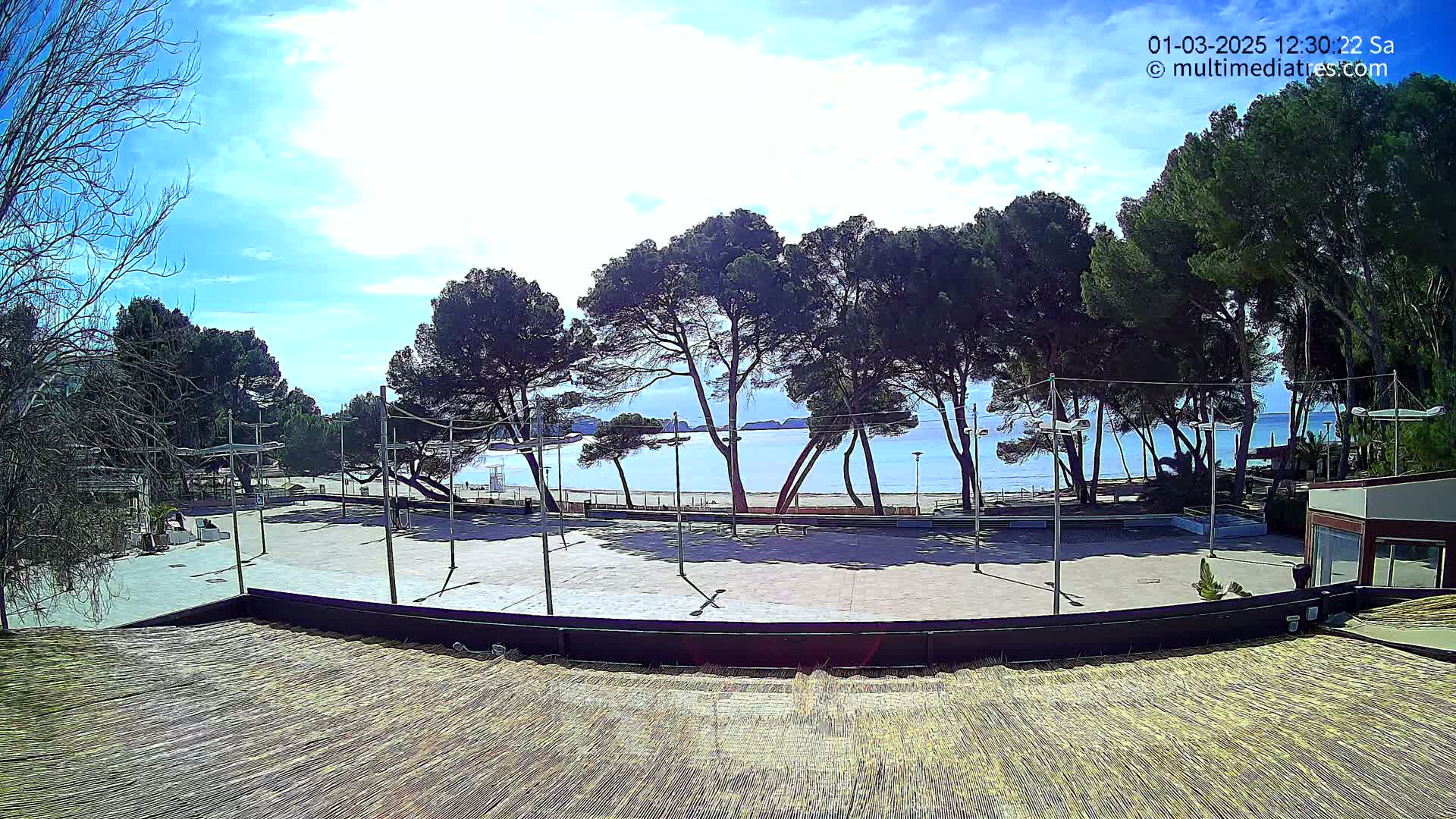 A mostly empty paved plaza overlooks a sandy beach and calm ocean under a mostly sunny sky.