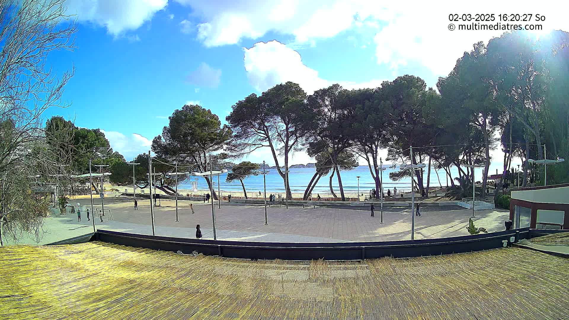 A paved plaza with a few people, overlooking a sandy beach and calm ocean under a mostly sunny sky.