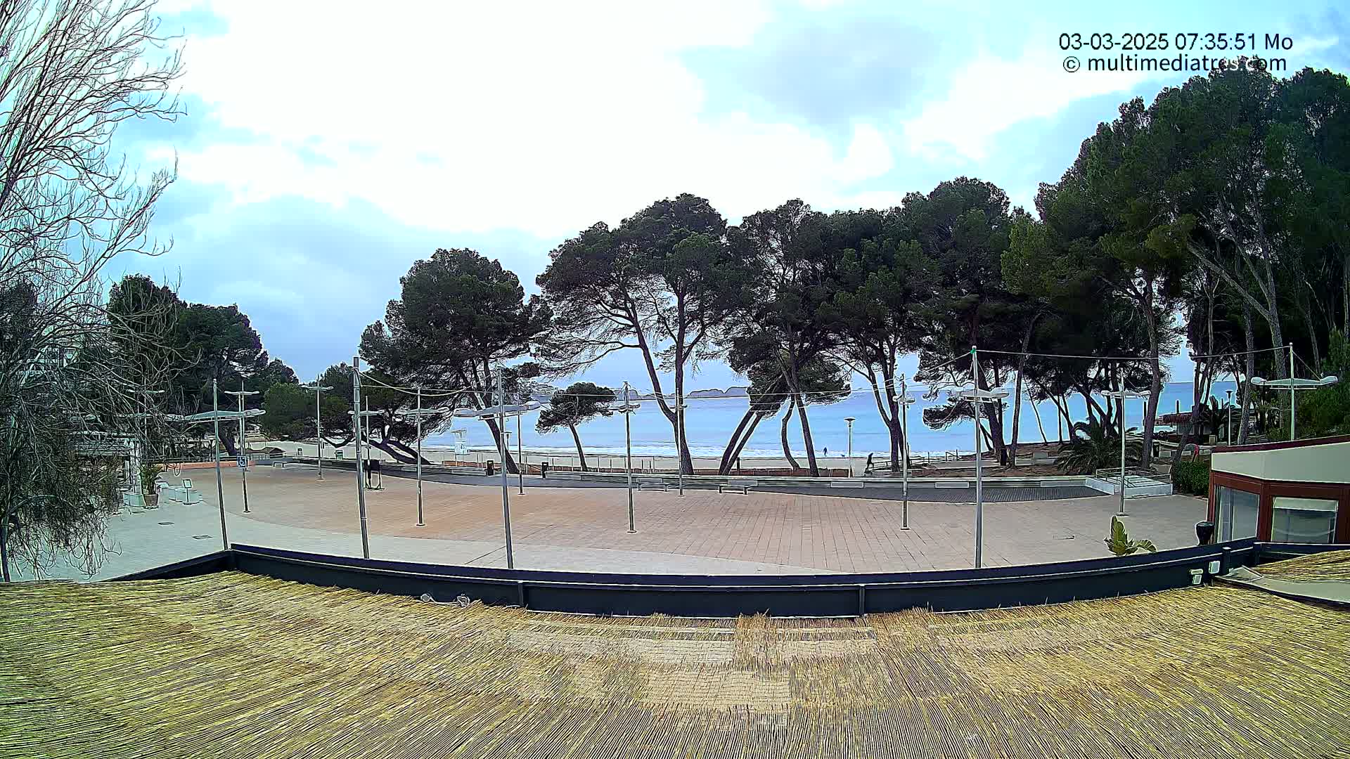 A paved plaza overlooks a sandy beach and calm ocean under a mostly cloudy sky, with tall pine trees lining the beach.
