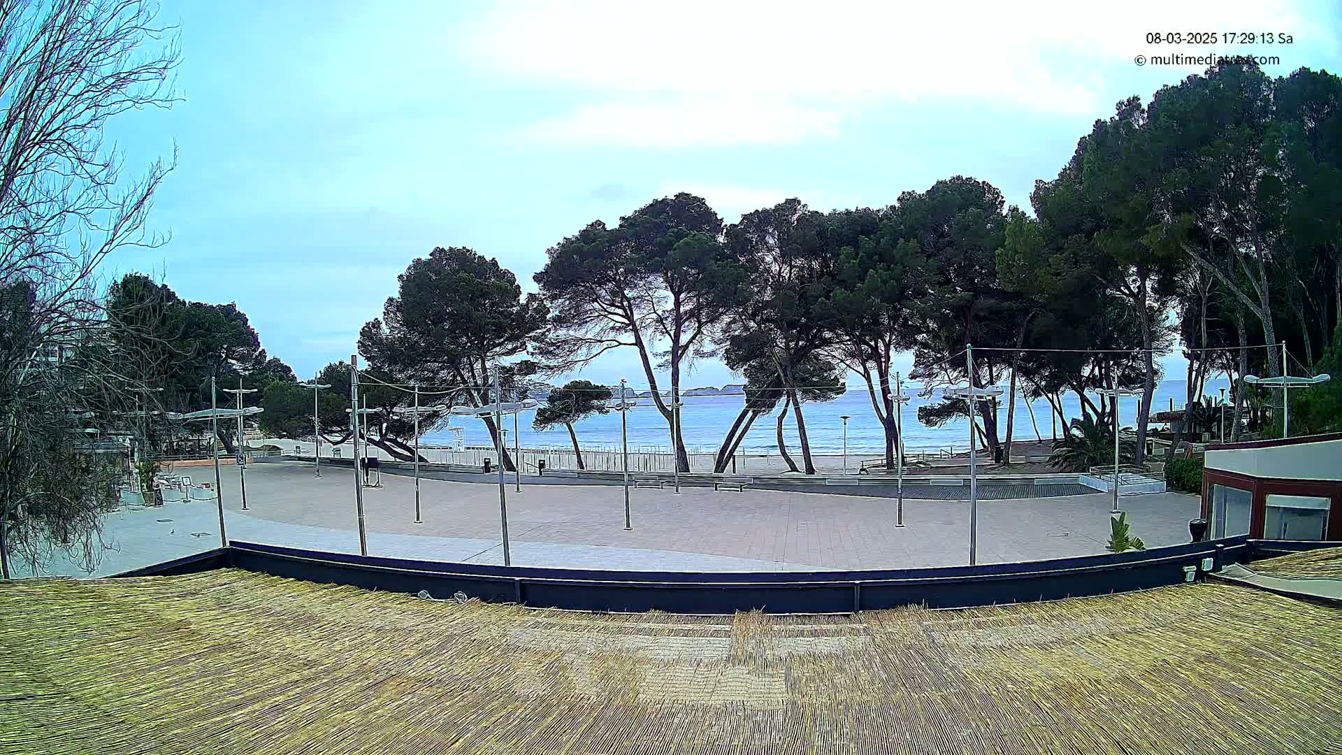 A mostly sunny day shows a paved plaza overlooking a beach with calm turquoise water and a row of pine trees, viewed from a thatched roof.