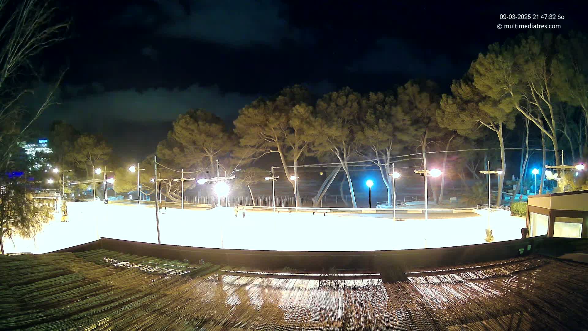 A brightly lit, empty plaza at night, surrounded by trees under a cloudy sky, is viewed from above a wet, slatted surface.
