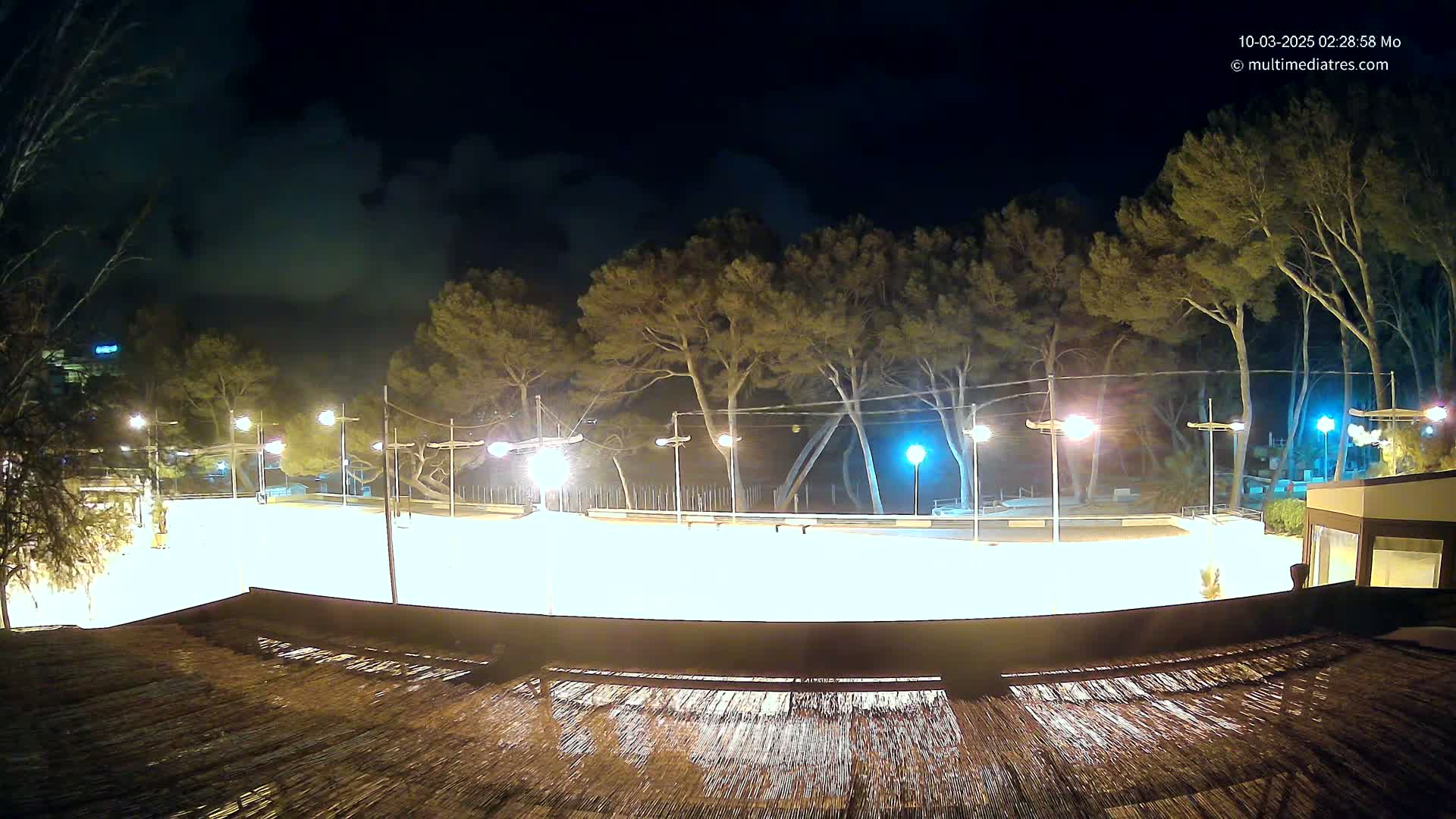 A brightly lit, empty outdoor rink at night, surrounded by trees under a cloudy sky.