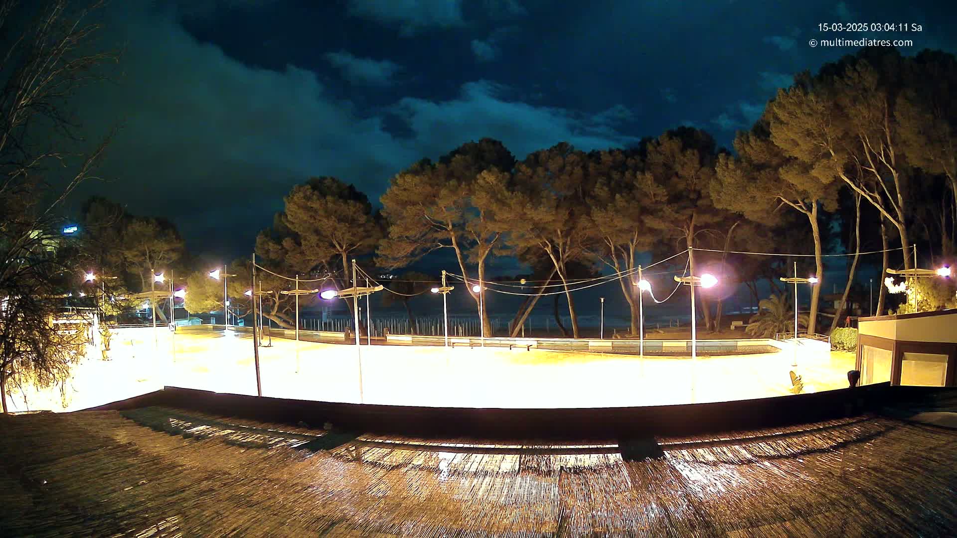 A brightly lit, empty, outdoor rink sits beneath a partly cloudy night sky next to a line of trees and a beach.