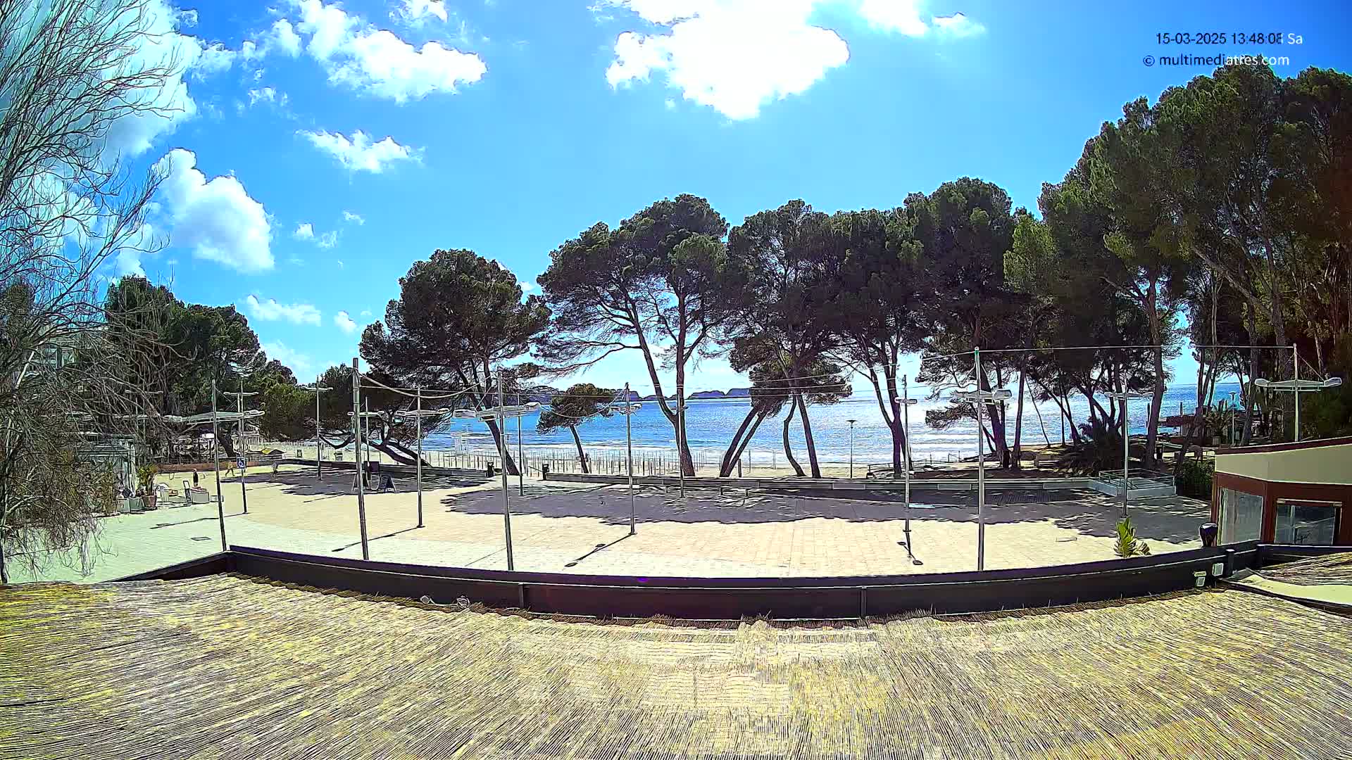 A paved plaza overlooks a sandy beach and calm blue ocean under a partly cloudy sky.