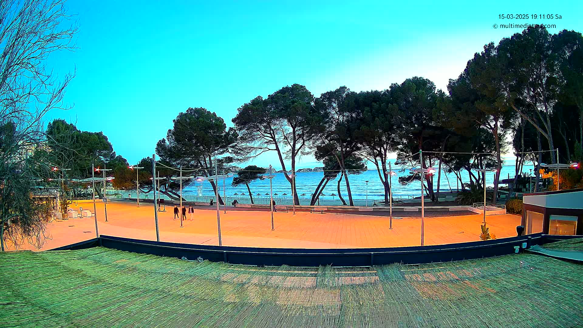 A paved plaza overlooks a calm ocean beach at dusk, lined with trees and illuminated by streetlights.