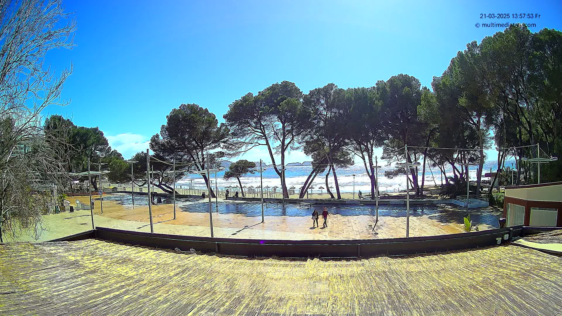 A paved plaza with a couple walking, overlooks a beach with a somewhat wavy ocean under a clear blue sky.