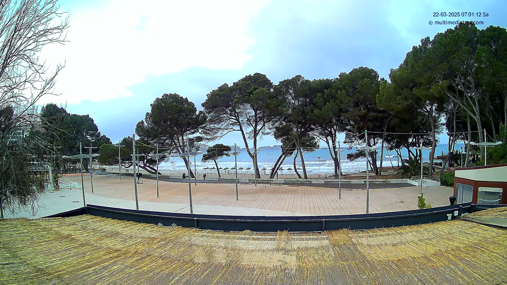 A paved plaza with a thatched roof structure overlooks a beach with gently rolling waves under a mostly cloudy sky, lined with pine trees.