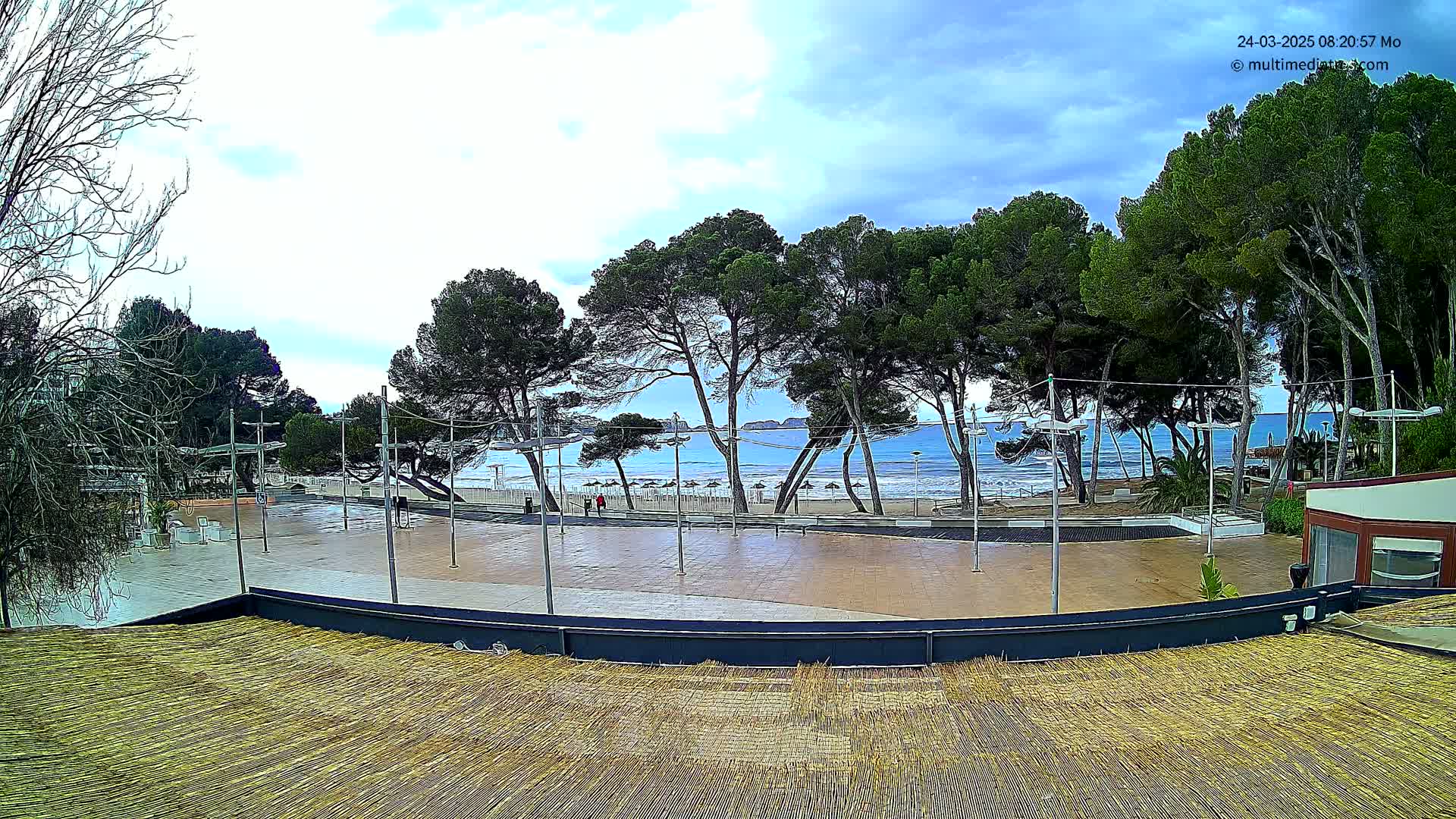 A paved plaza overlooks a beach with calm, blue water under a partly cloudy sky, lined with pine trees.