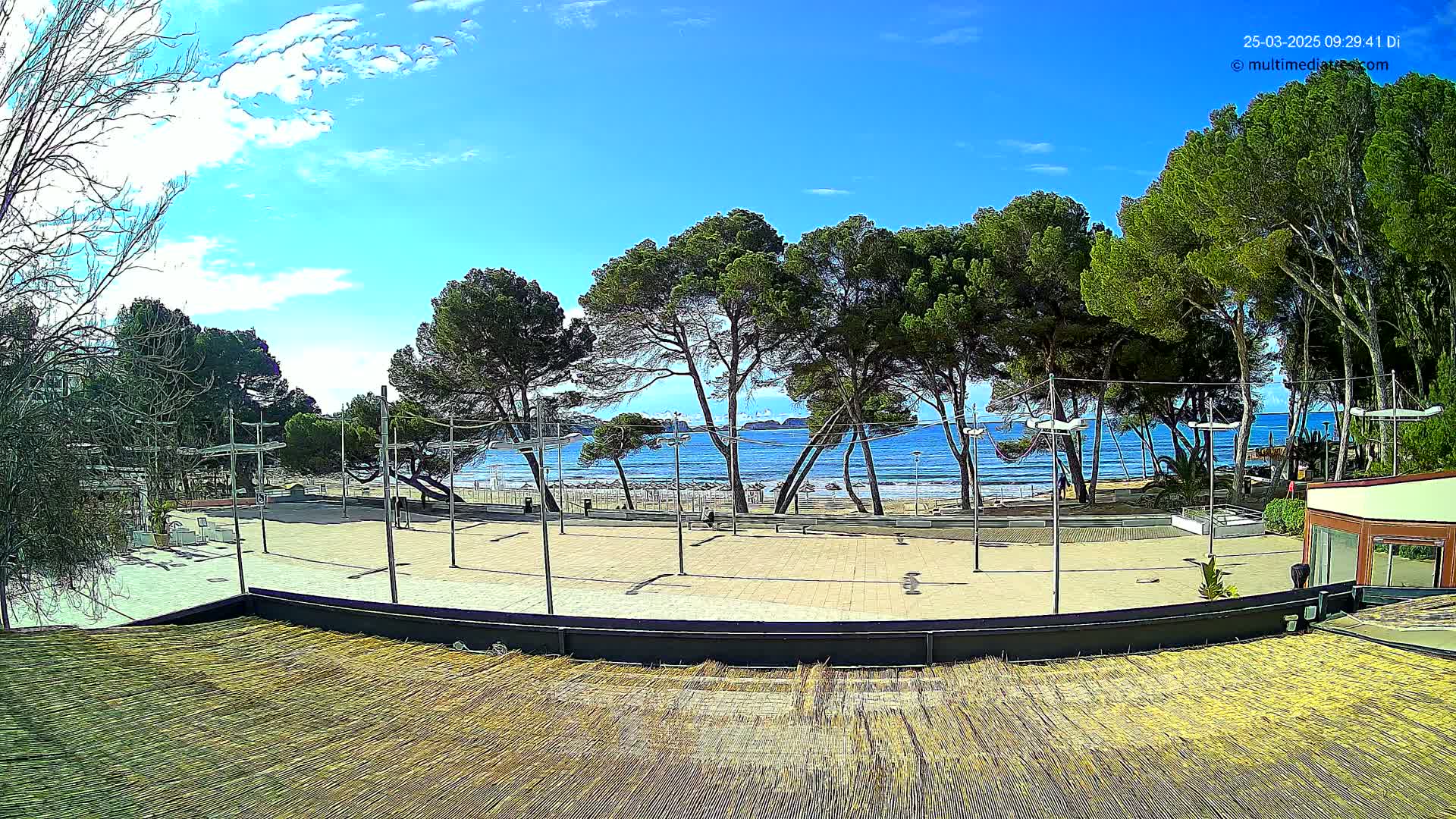 A paved plaza overlooks a sandy beach and calm blue ocean under a mostly sunny sky.
