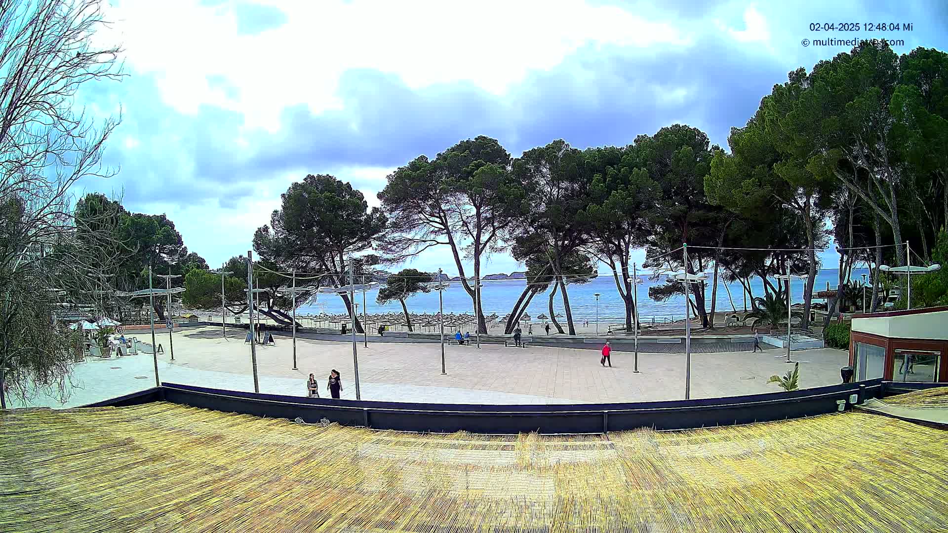 A partly cloudy day overlooks a beachfront plaza with a few people walking, lined with tall pine trees and a sandy beach with calm water.