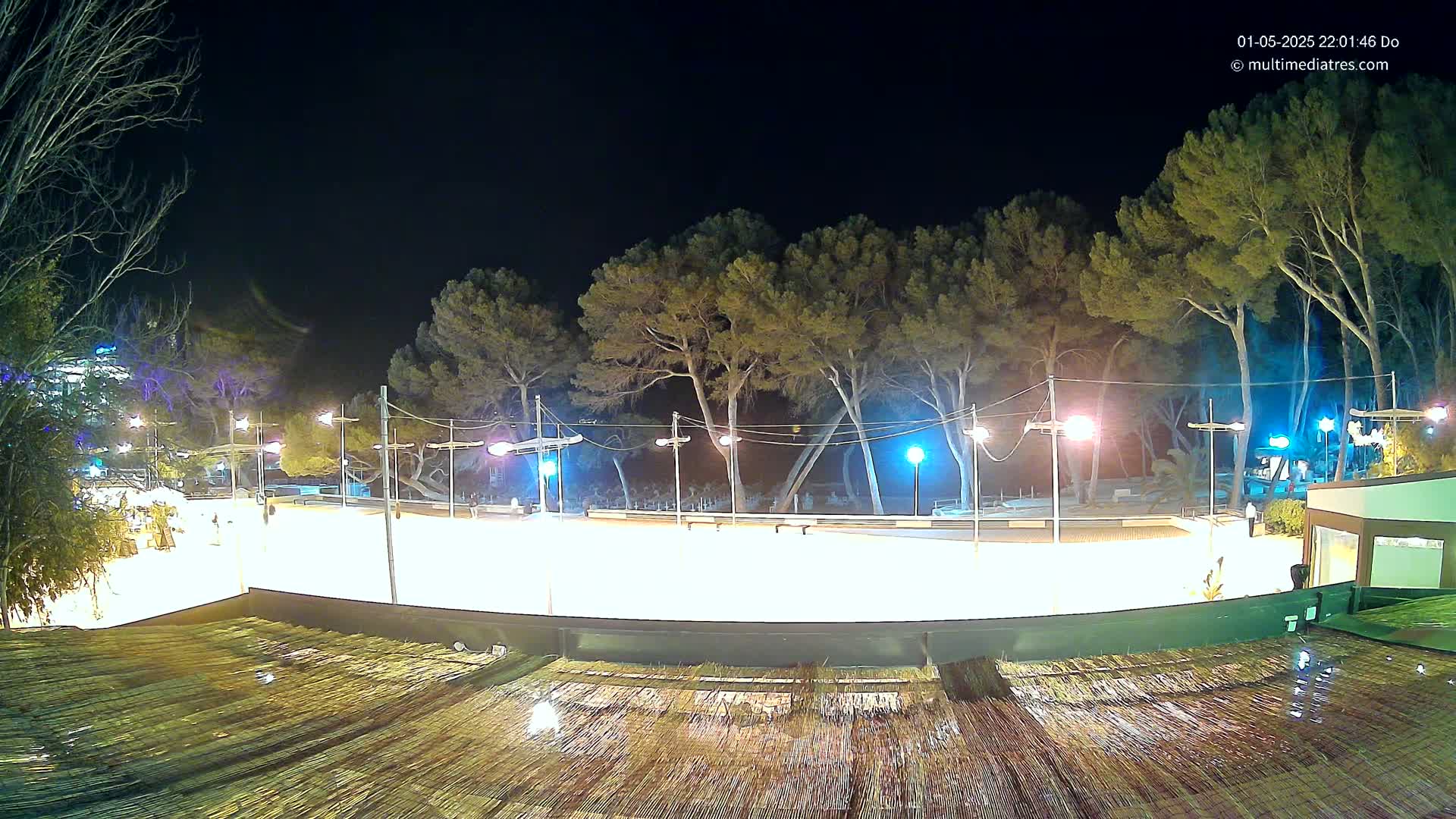 A brightly lit outdoor ice rink at night, surrounded by trees and illuminated by streetlights, with a wet thatched roof in the foreground.