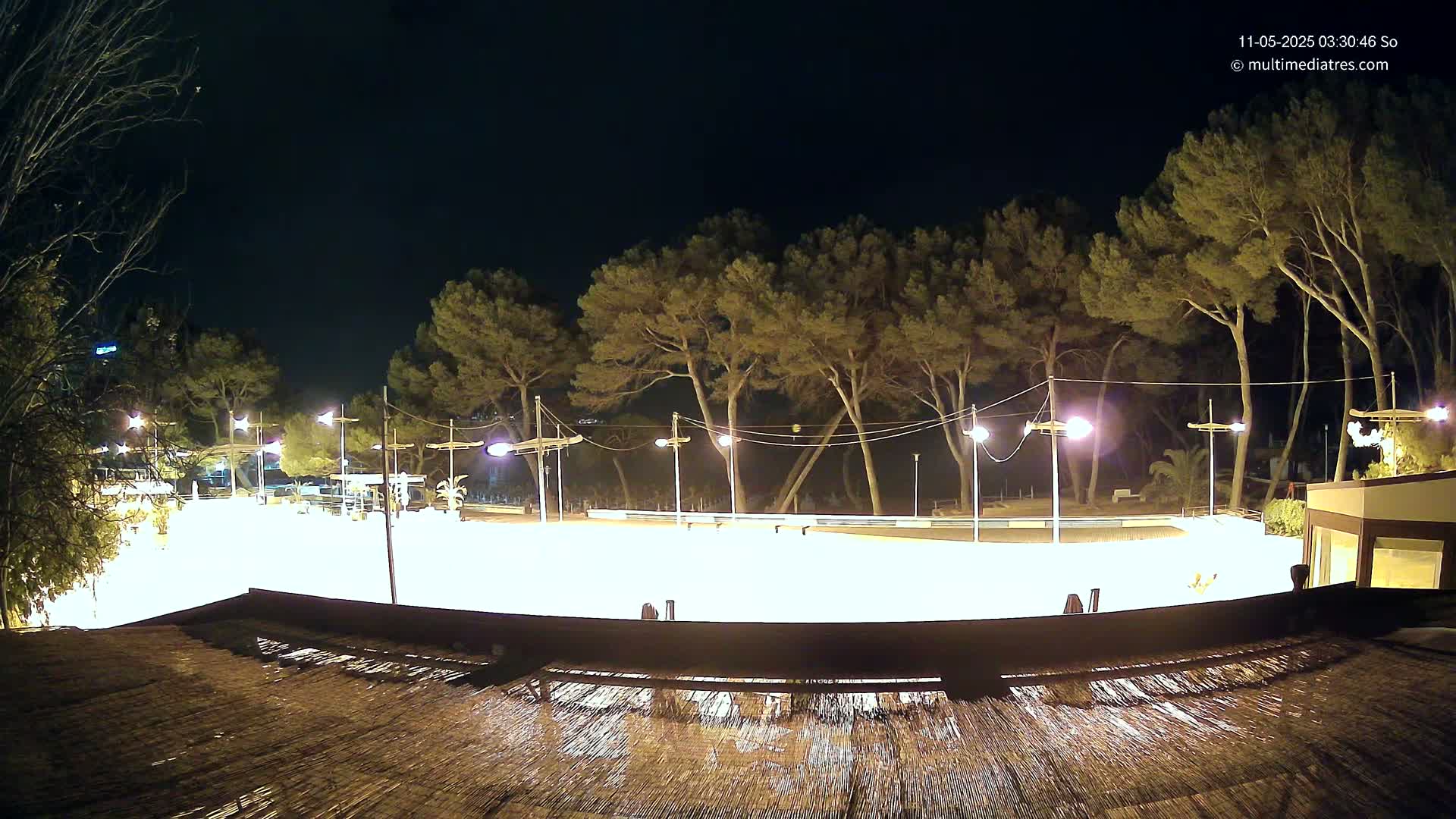 A brightly lit, empty plaza at night, surrounded by trees and illuminated by lampposts, under a clear, dark sky.