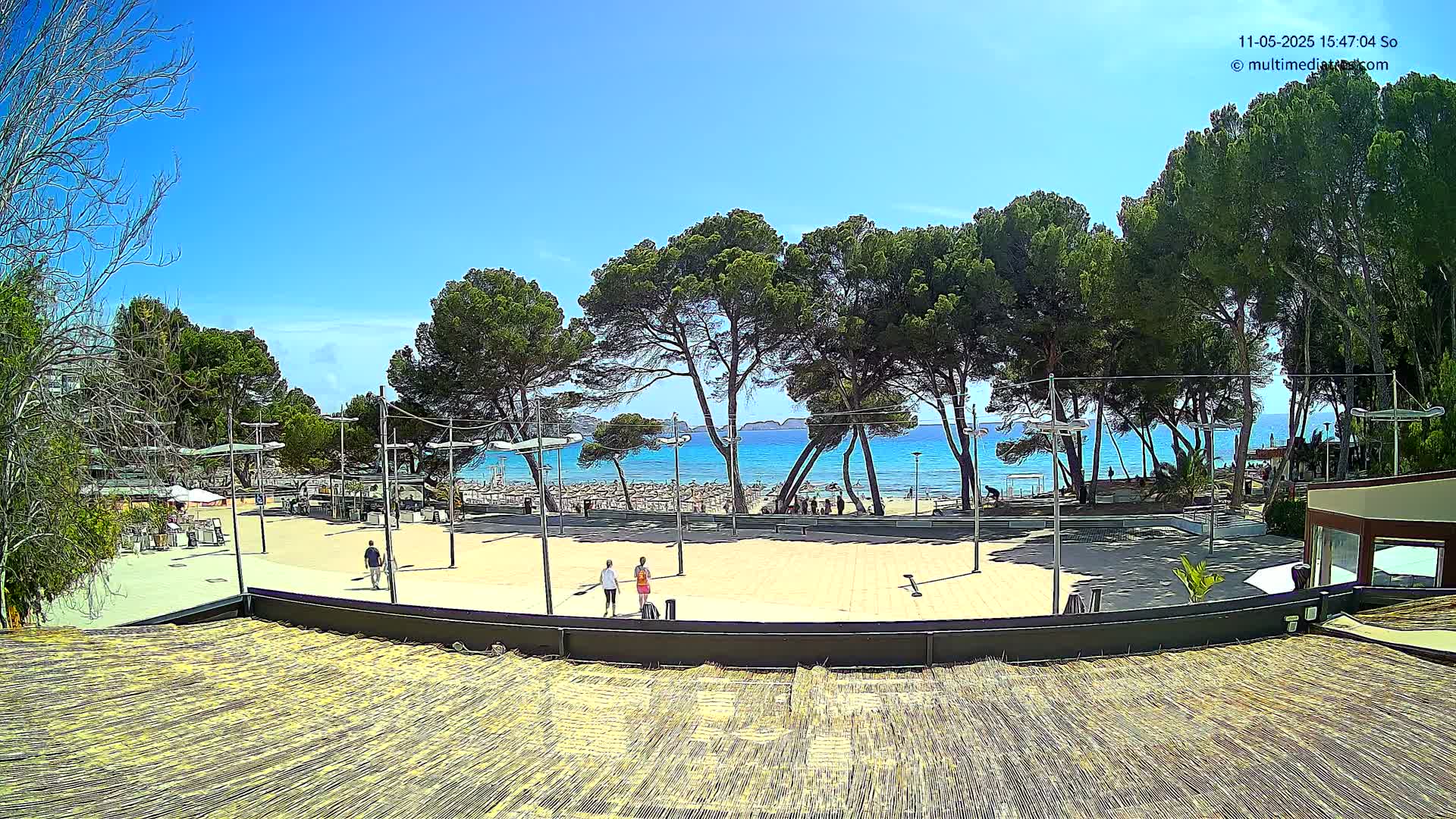 A paved plaza with a few people walking overlooks a sandy beach with turquoise water under a clear blue sky.