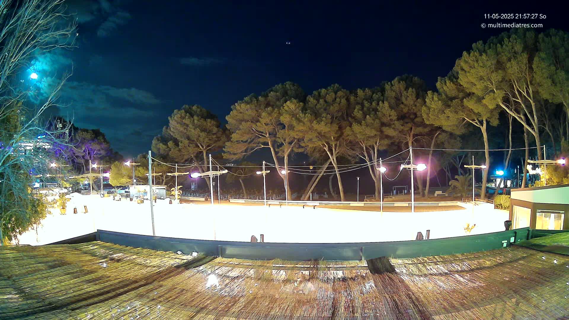 A brightly lit, empty outdoor rink is surrounded by trees under a partly cloudy night sky.
