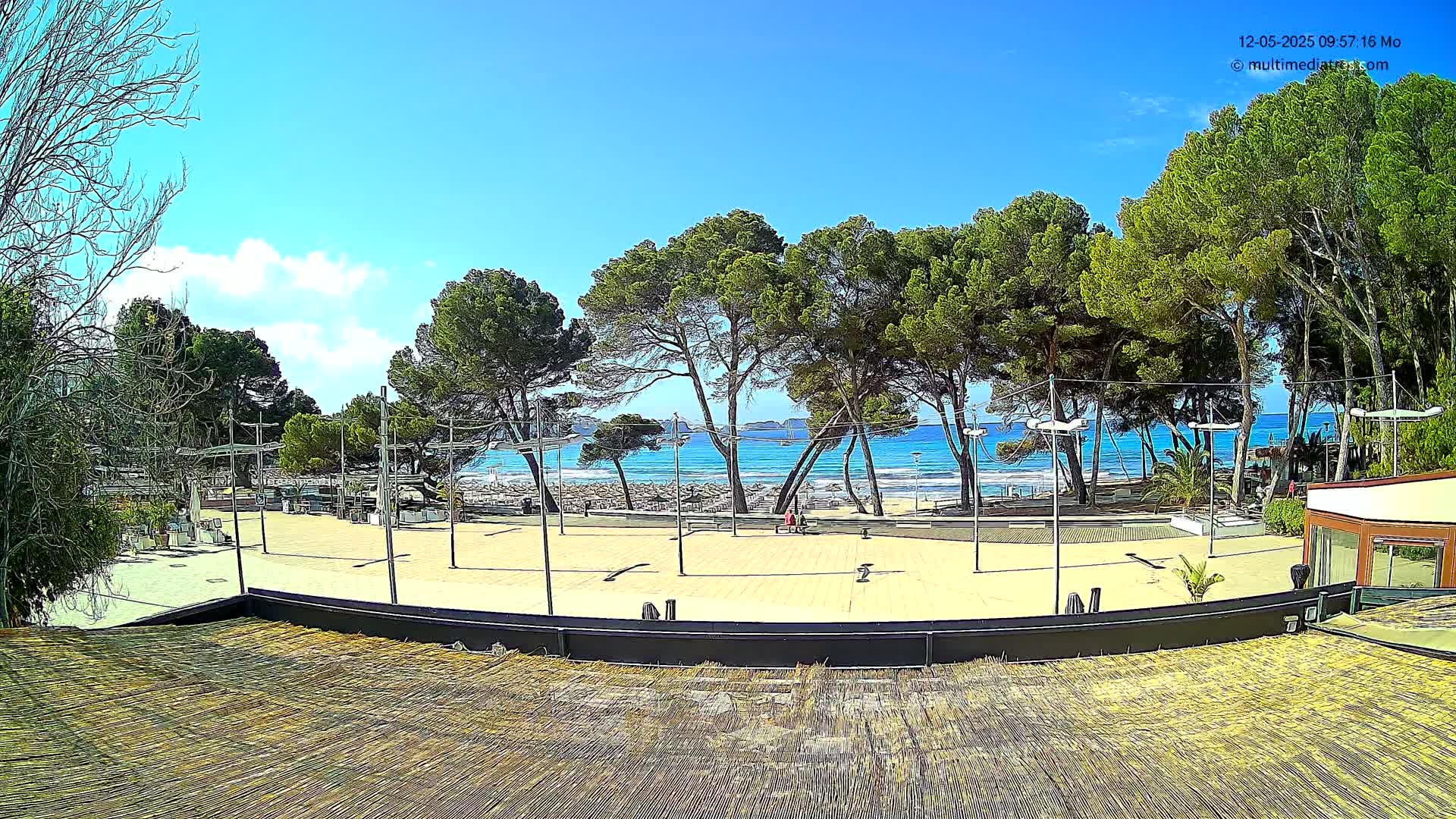 A sandy plaza overlooks a beach with turquoise water under a mostly sunny sky, lined with pine trees.