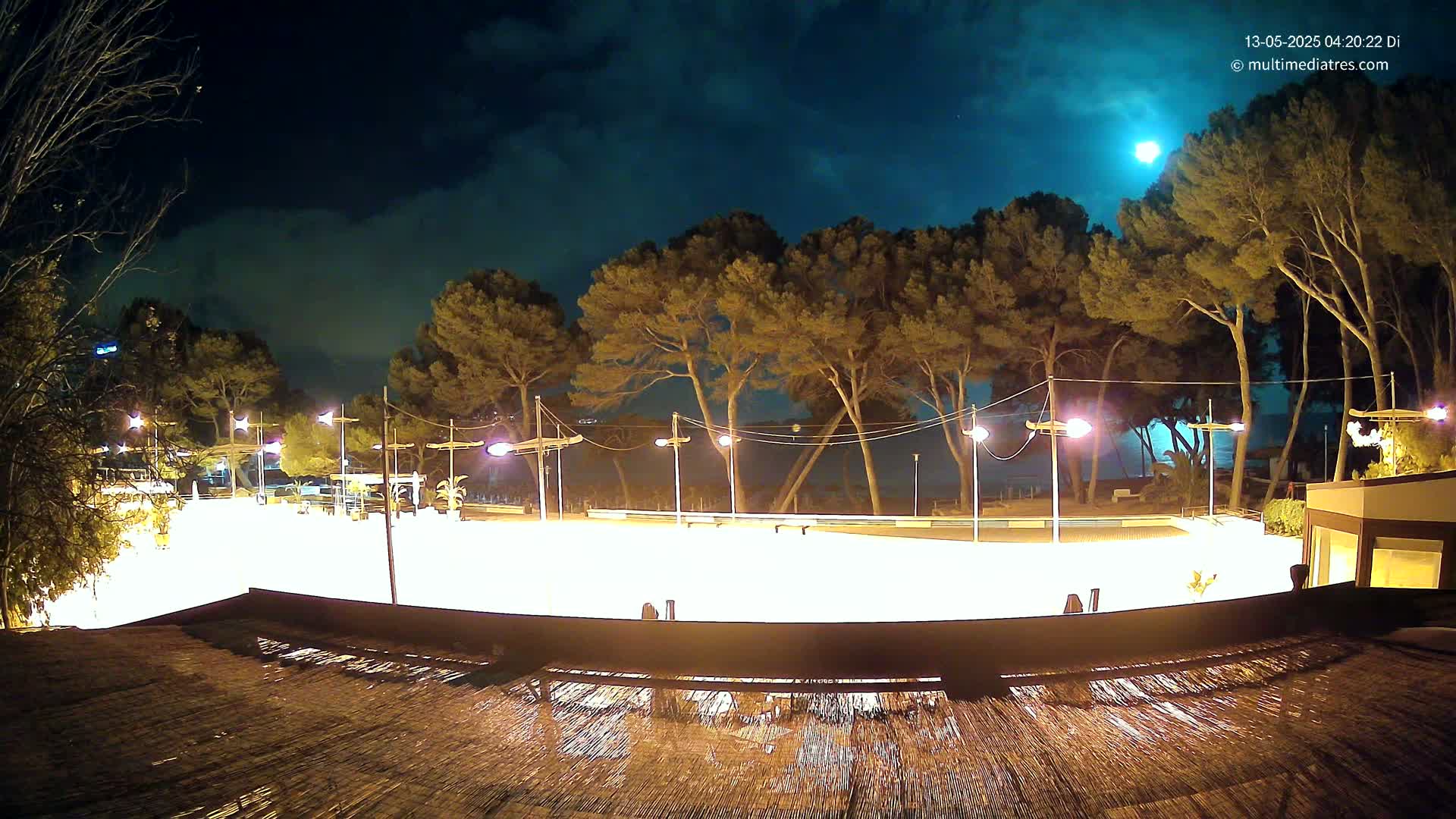 A brightly lit, empty plaza at night, under a partly cloudy sky with a visible moon, is surrounded by tall, dark trees near a body of water.