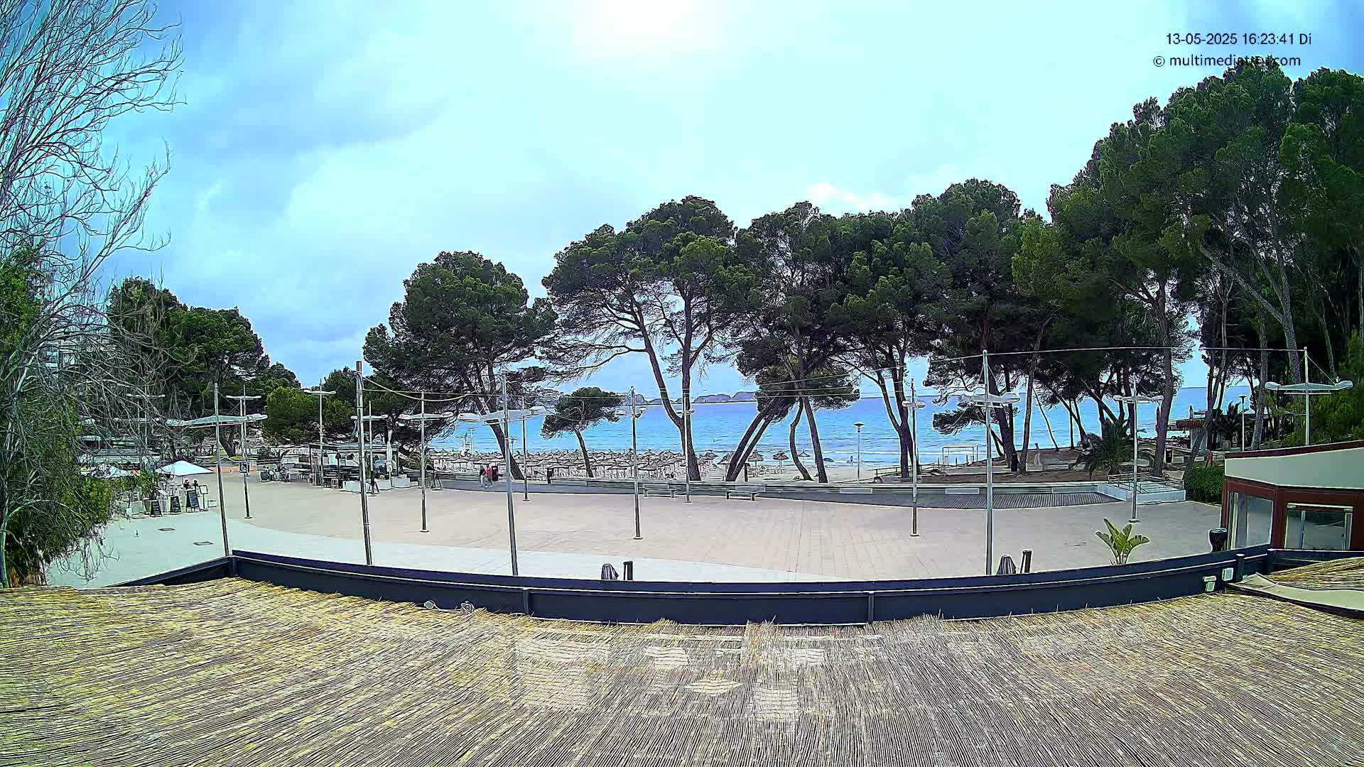 A mostly sunny day reveals a paved plaza overlooking a beach with calm, blue water and a line of pine trees.