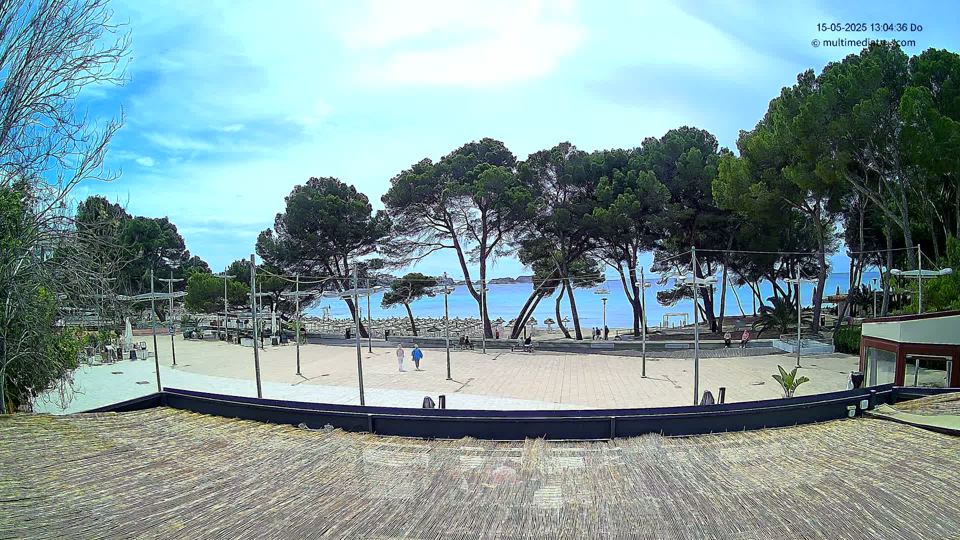 A paved plaza with several people walking, overlooks a calm, blue ocean beach lined with trees under a partly cloudy sky.