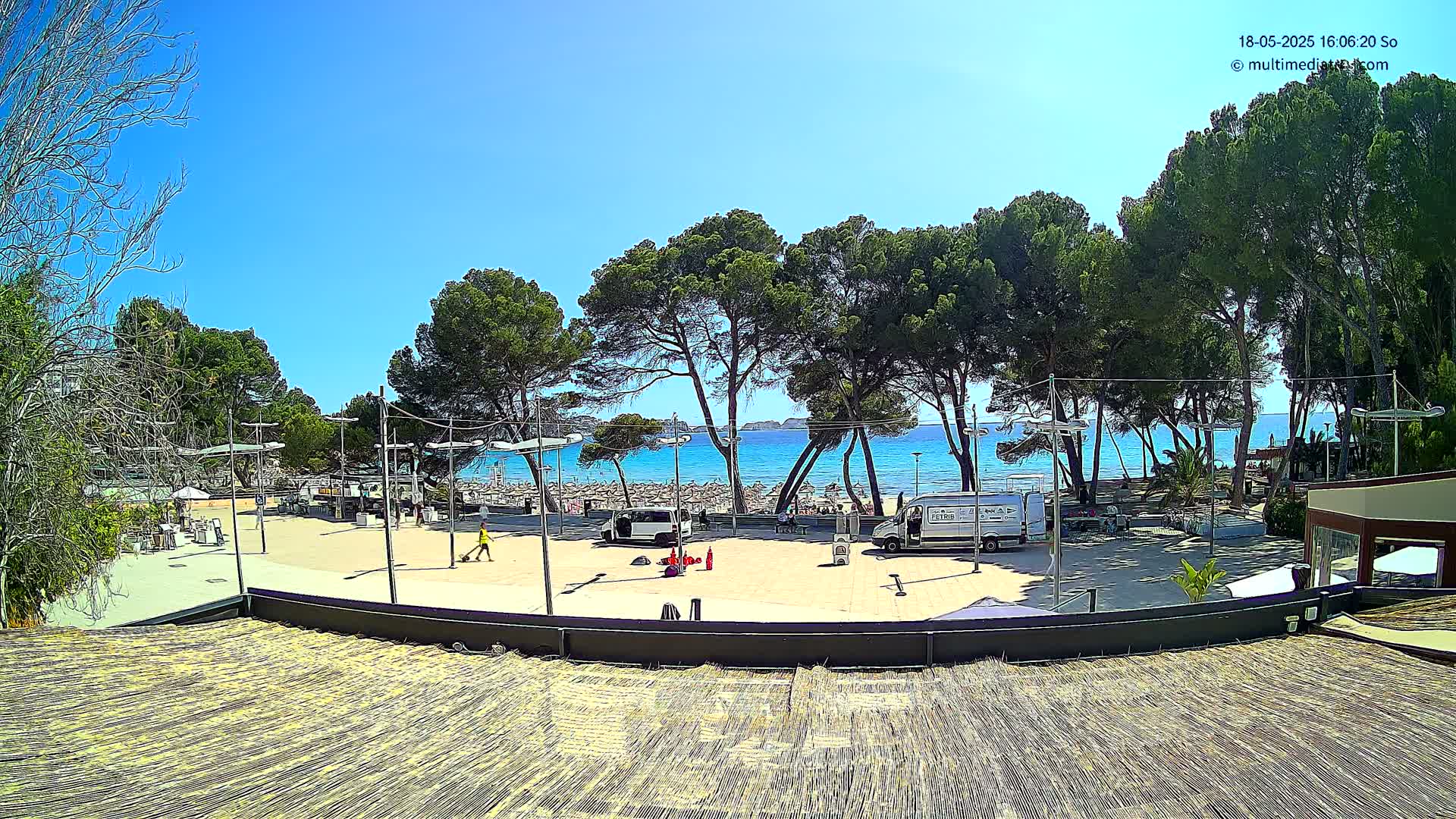 A sunny beach scene is viewed from an elevated position, showing a paved plaza with a few people and vehicles, surrounded by pine trees, leading to a sandy beach and calm blue water.