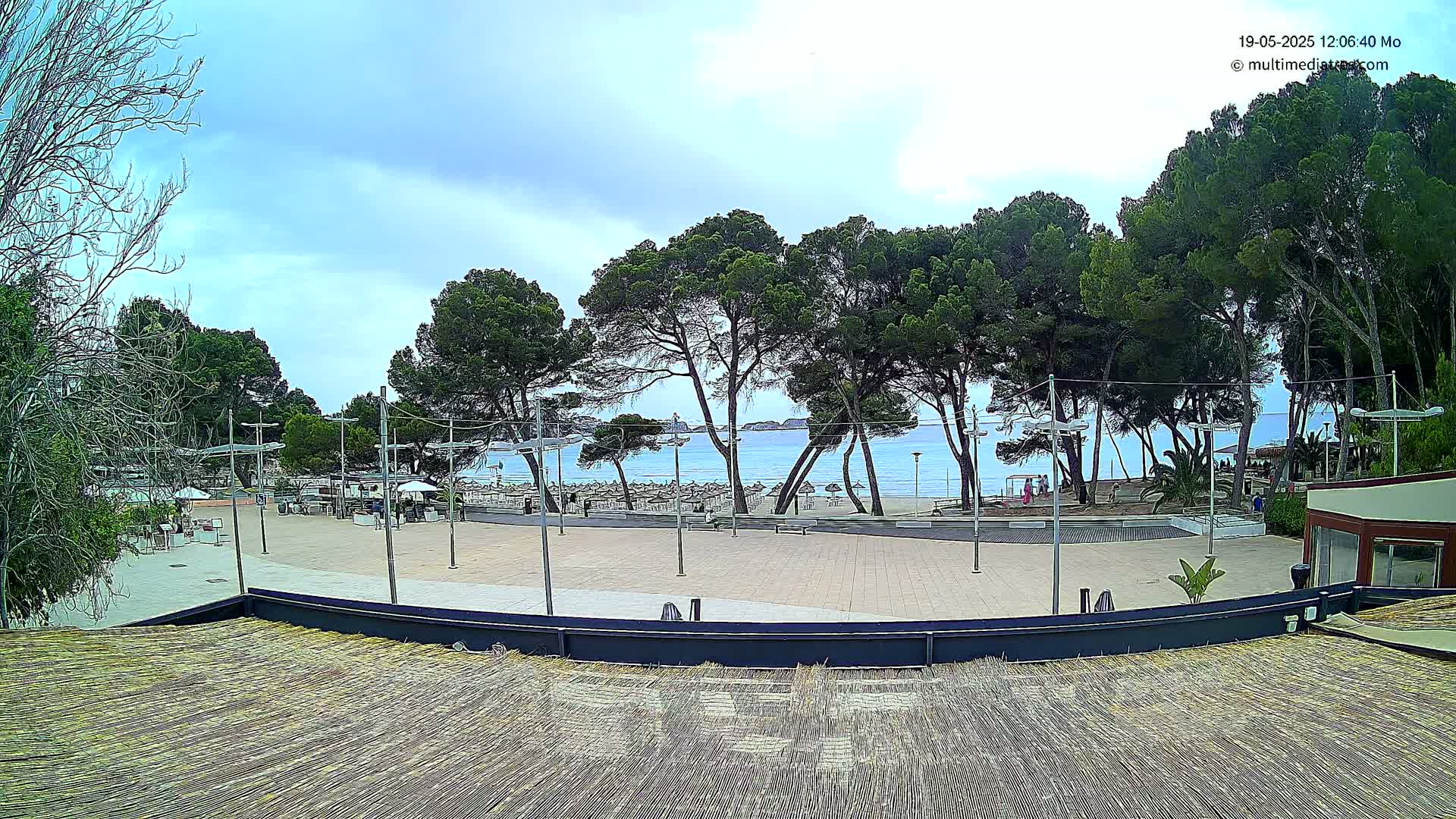 A view from a rooftop shows a paved plaza overlooking a beach with calm water under a partly cloudy sky, surrounded by tall pine trees.