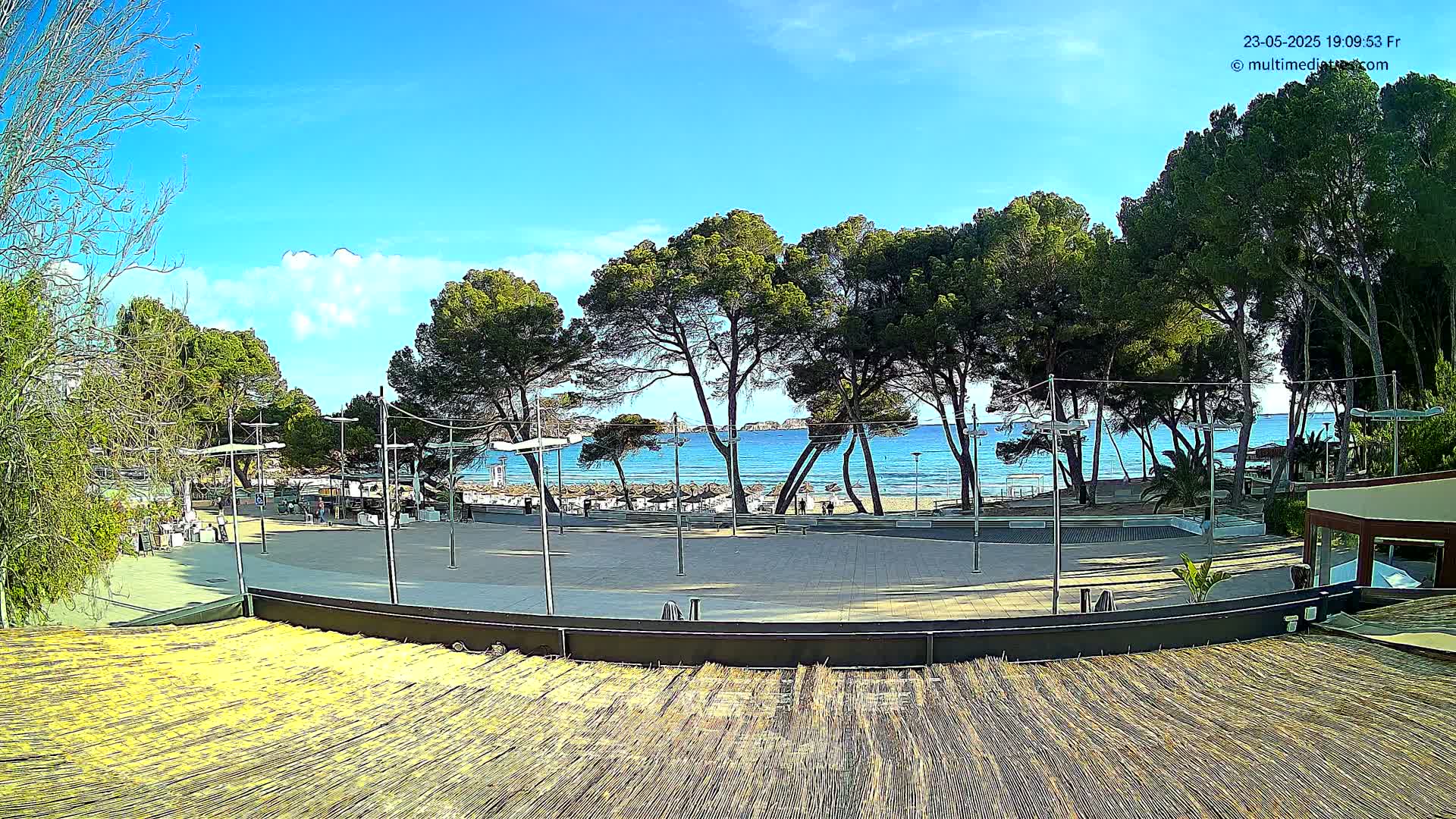 A paved plaza with a thatched roof structure overlooks a sandy beach and calm blue ocean under a mostly sunny sky.