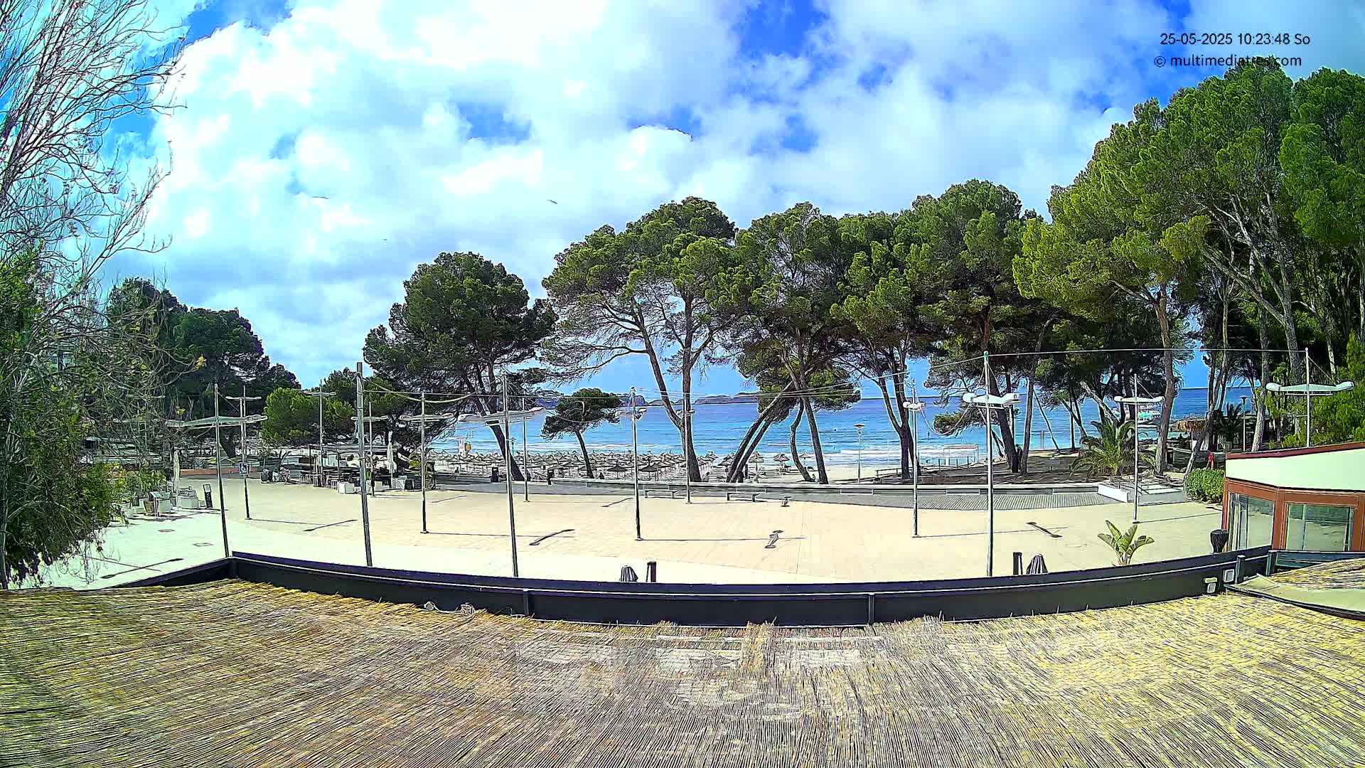 A paved plaza overlooks a sandy beach with turquoise water under a partly cloudy sky,  surrounded by lush green pine trees.