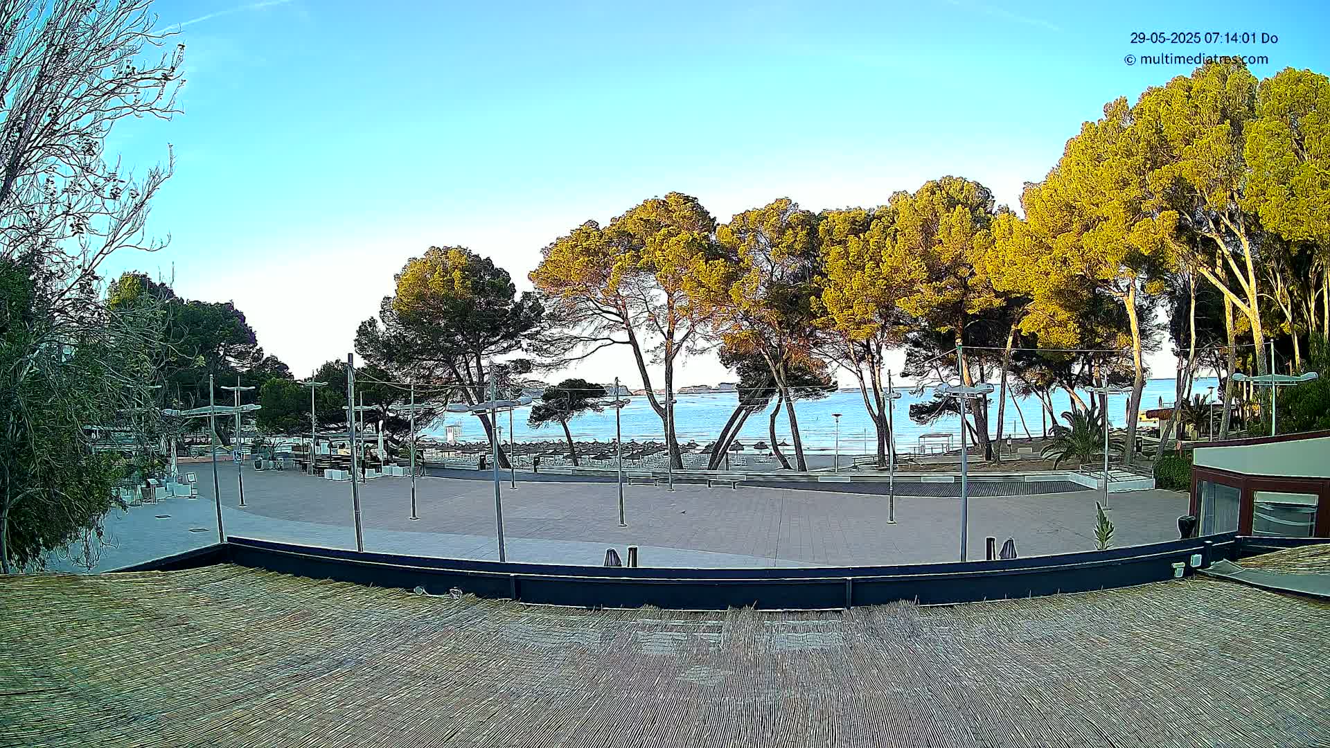 A paved plaza with a beach and calm ocean in the background under a clear, mostly sunny sky.