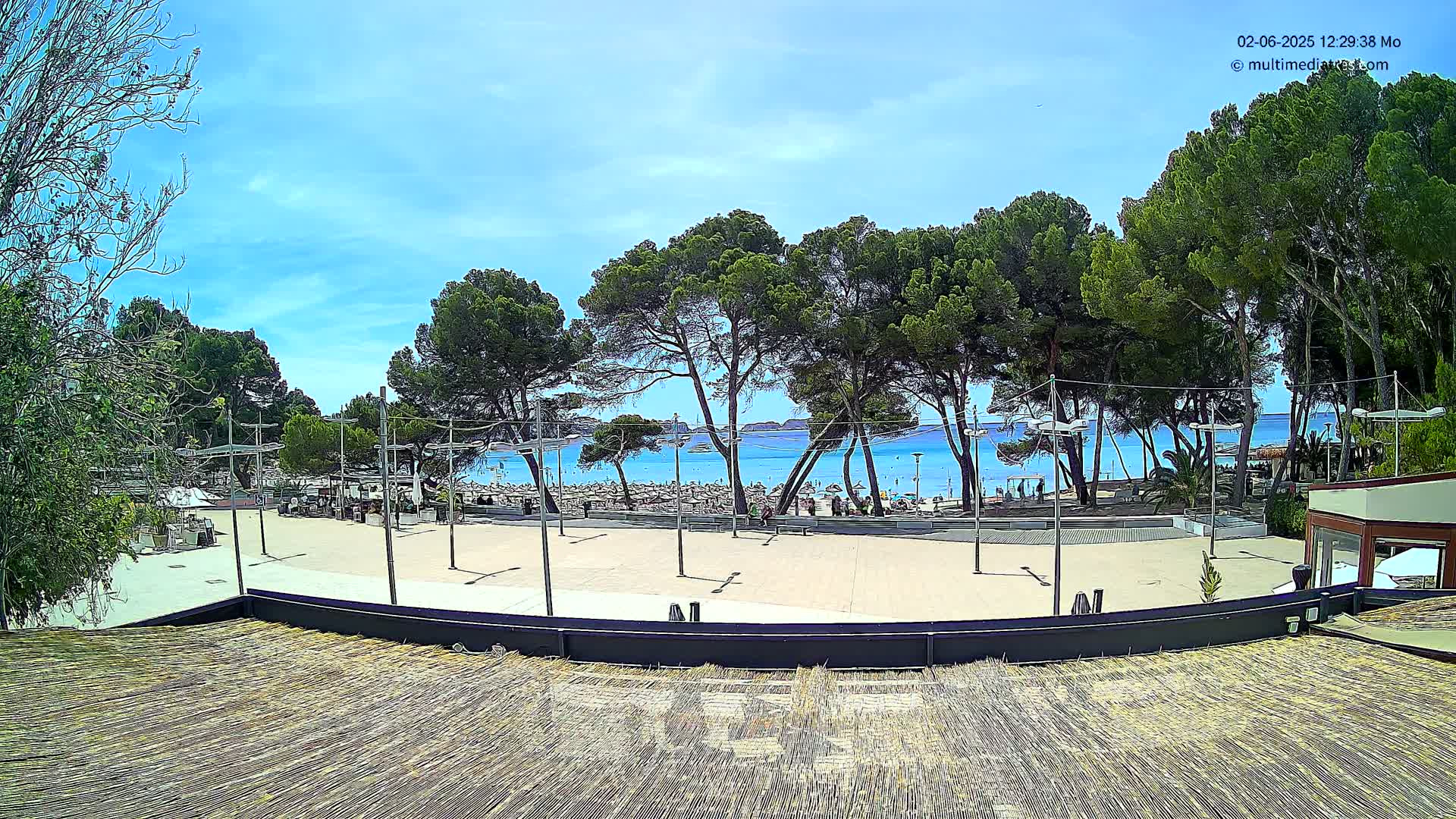 A sunny beach scene is visible from a high vantage point, showing a plaza with several trees and a crowded beach beyond, under a mostly clear blue sky.