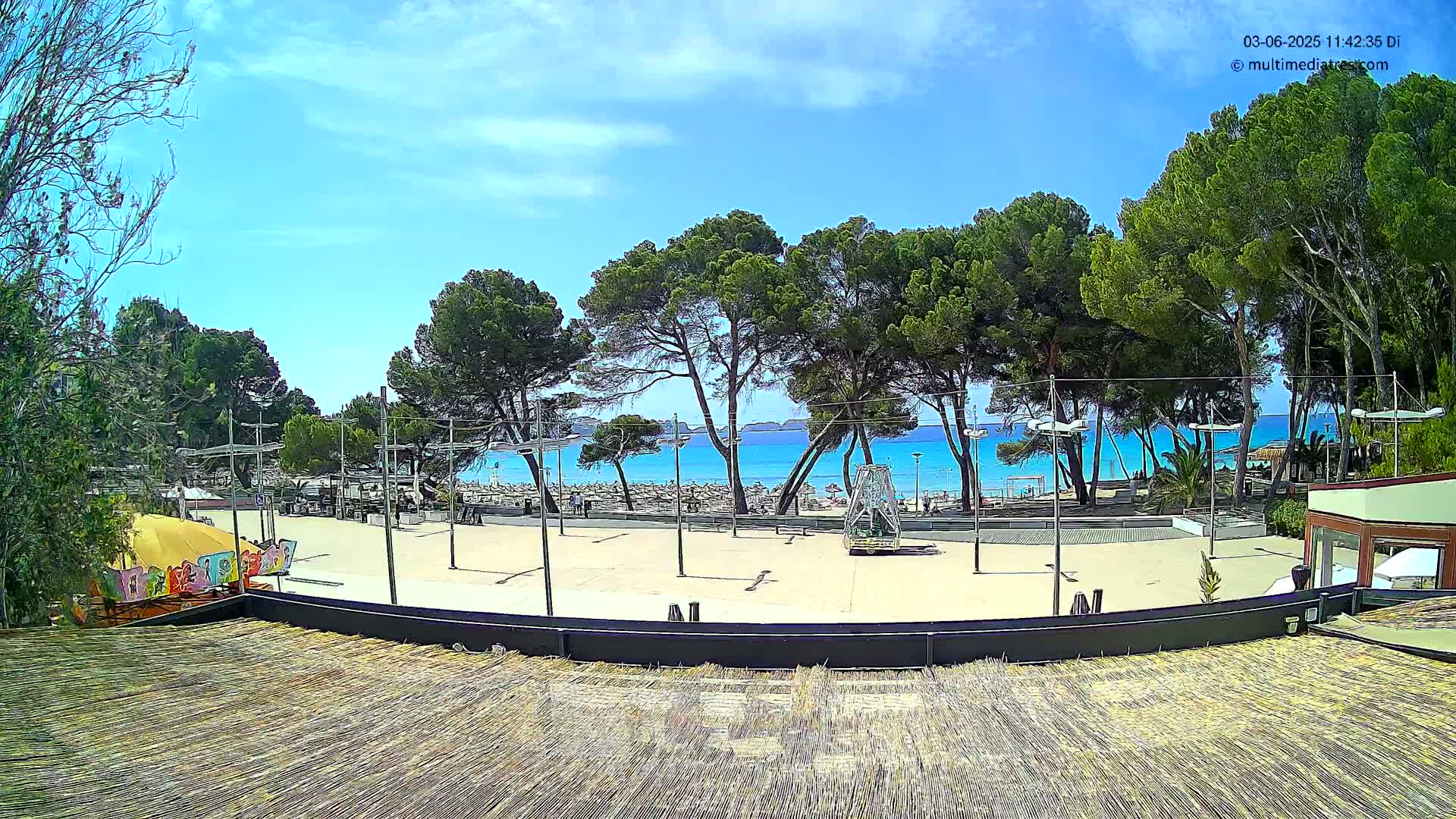 A sandy plaza with a beach and turquoise ocean visible beyond a row of pine trees under a mostly sunny sky.