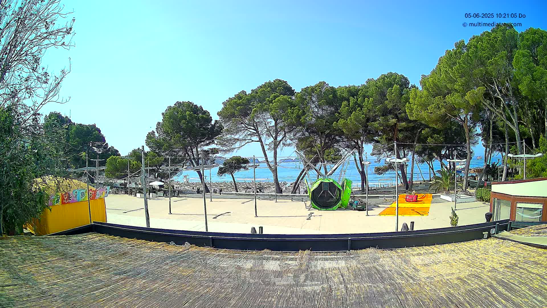 A sunny outdoor scene shows a beach with calm water visible through a line of pine trees, beyond which is a paved area with playground equipment, small buildings, and a yellow structure.