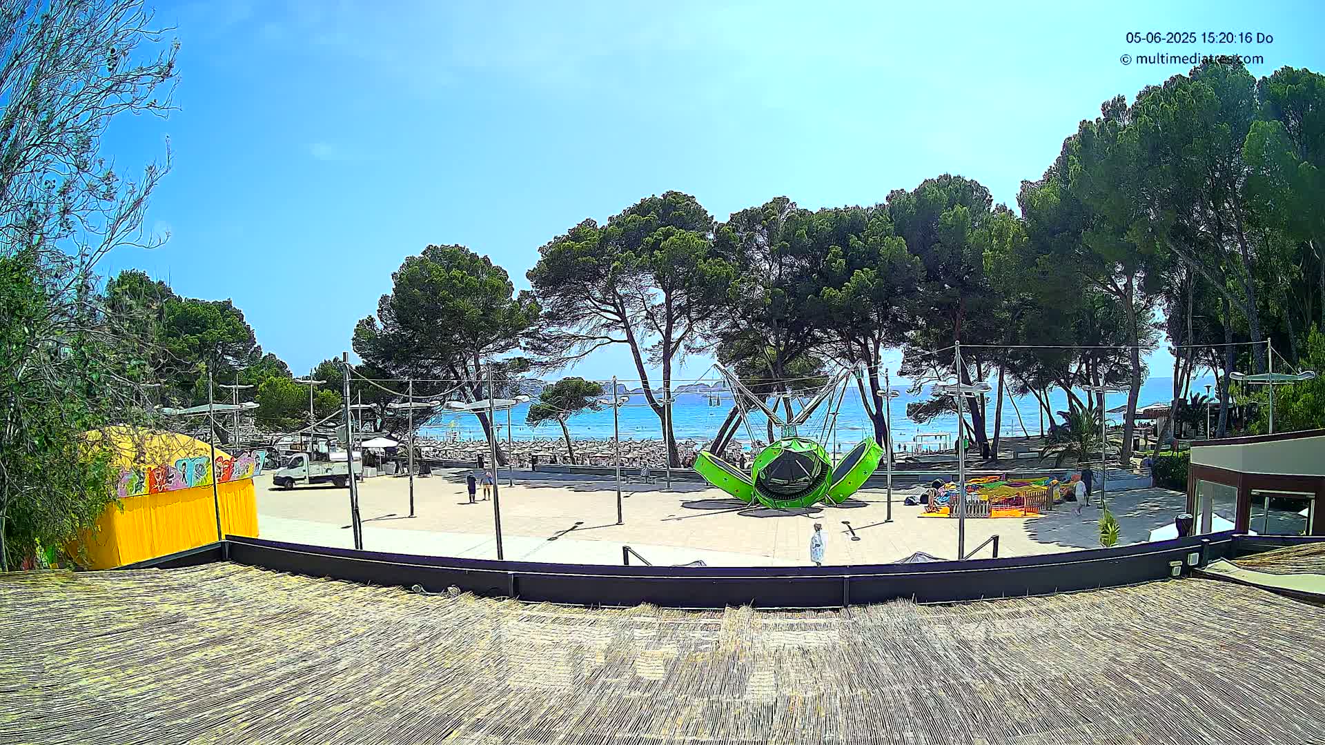 A sunny beach scene is visible beyond a paved area with a playground featuring a large green ride and smaller attractions, all situated among tall pine trees.