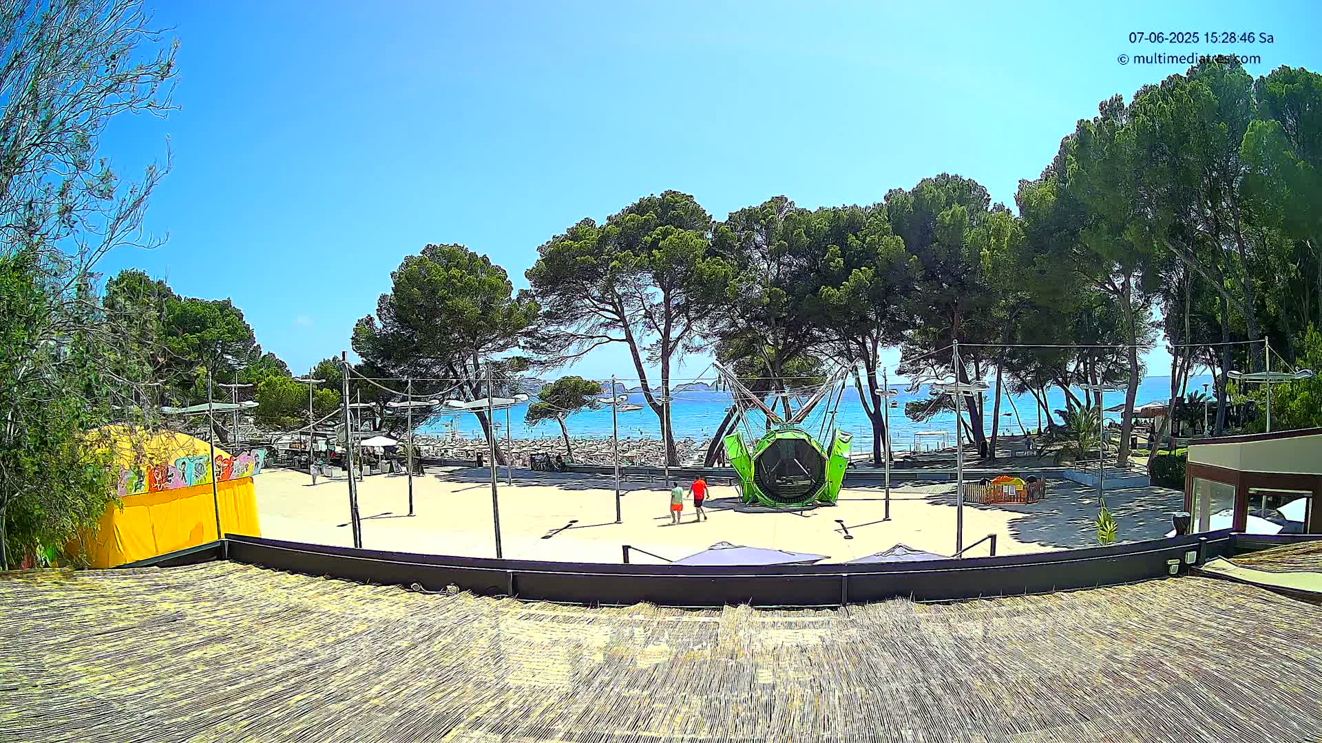 A sandy outdoor area with a playground, trees, and a beach with calm, clear blue water visible in the background under a sunny sky.