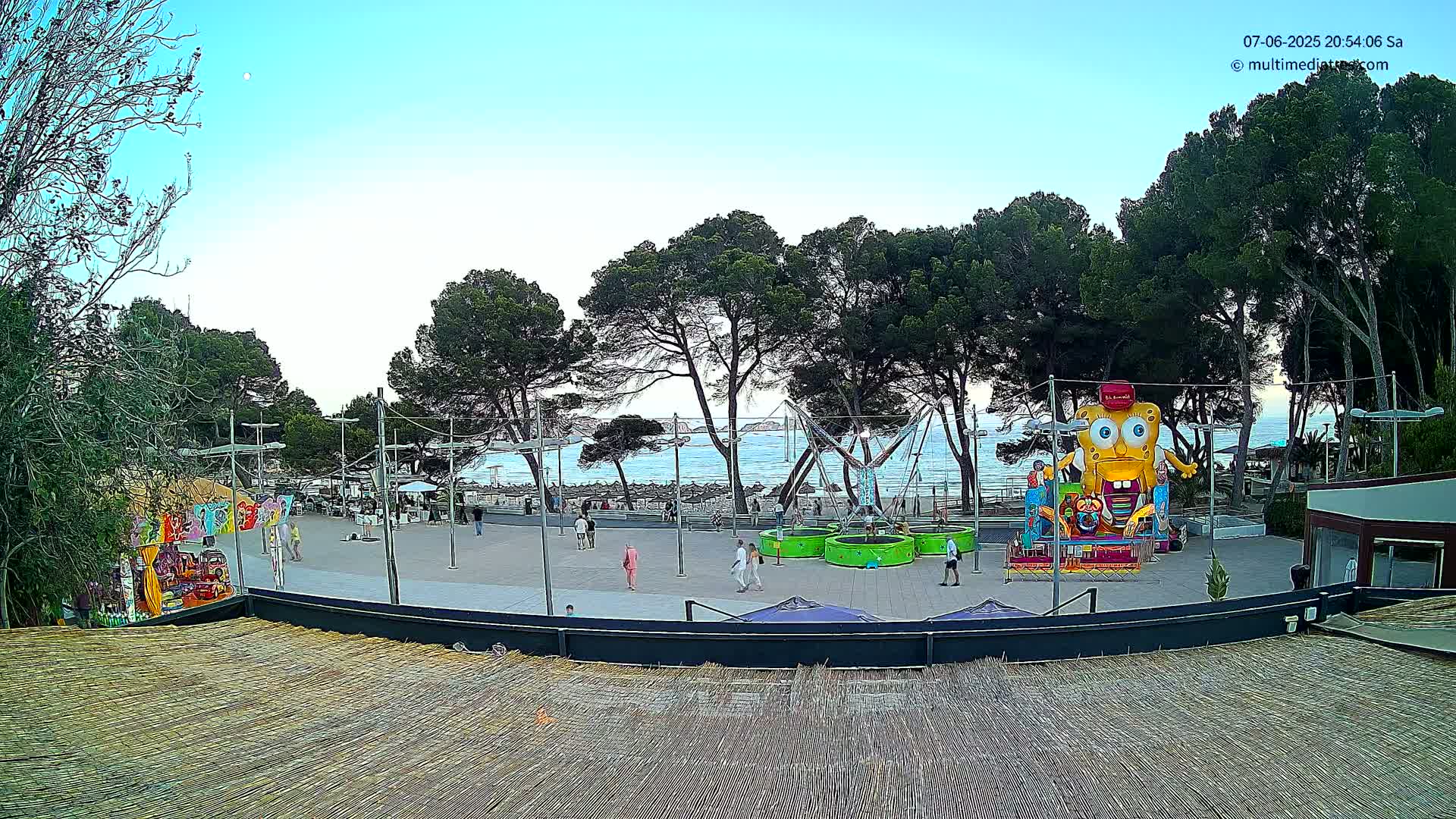 A seaside plaza with amusement park rides, including a large cartoon character inflatable, under a clear twilight sky.