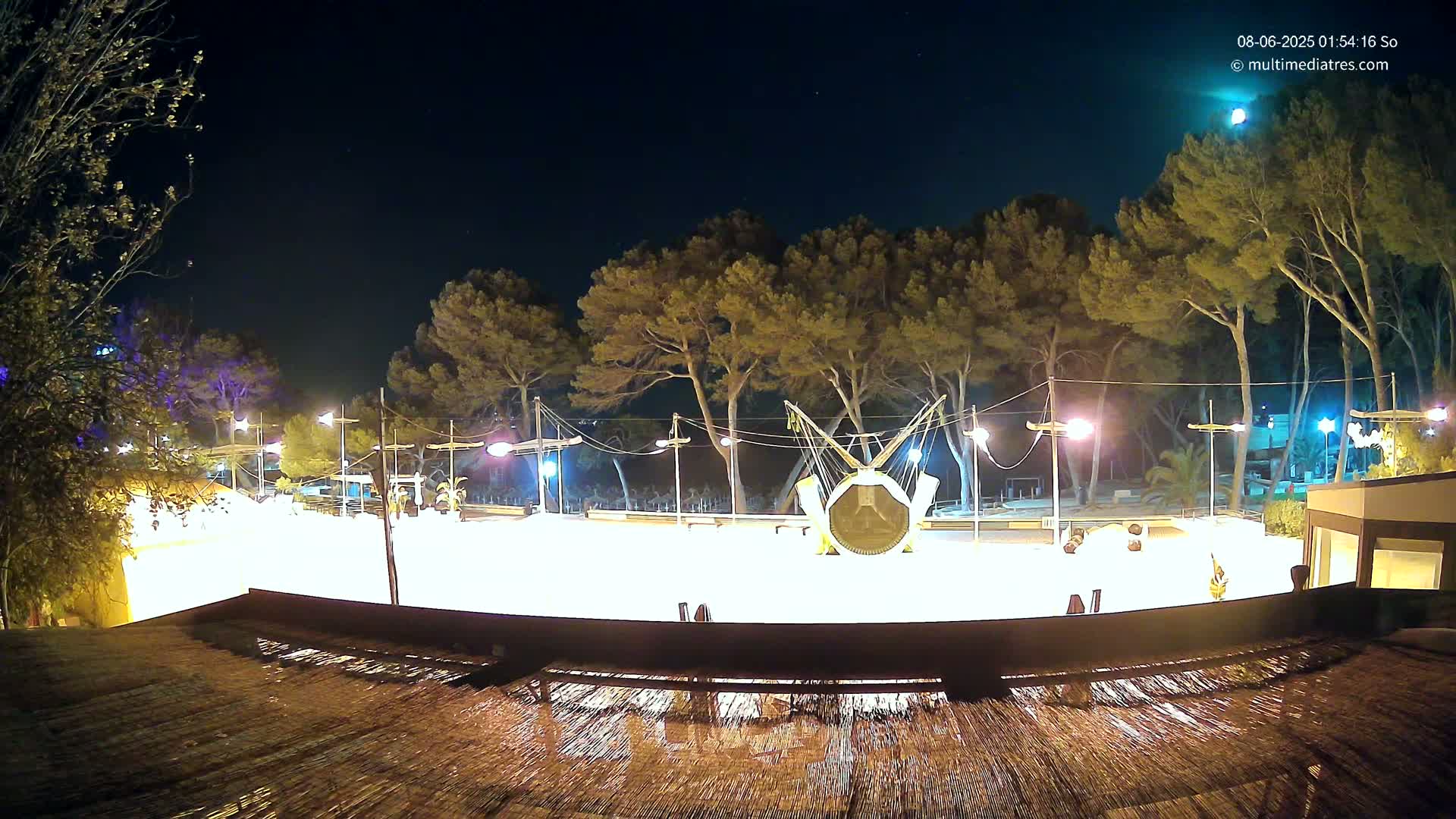A brightly lit outdoor area at night, featuring a trampoline-like structure surrounded by trees and illuminated by lampposts under a clear, starry sky.