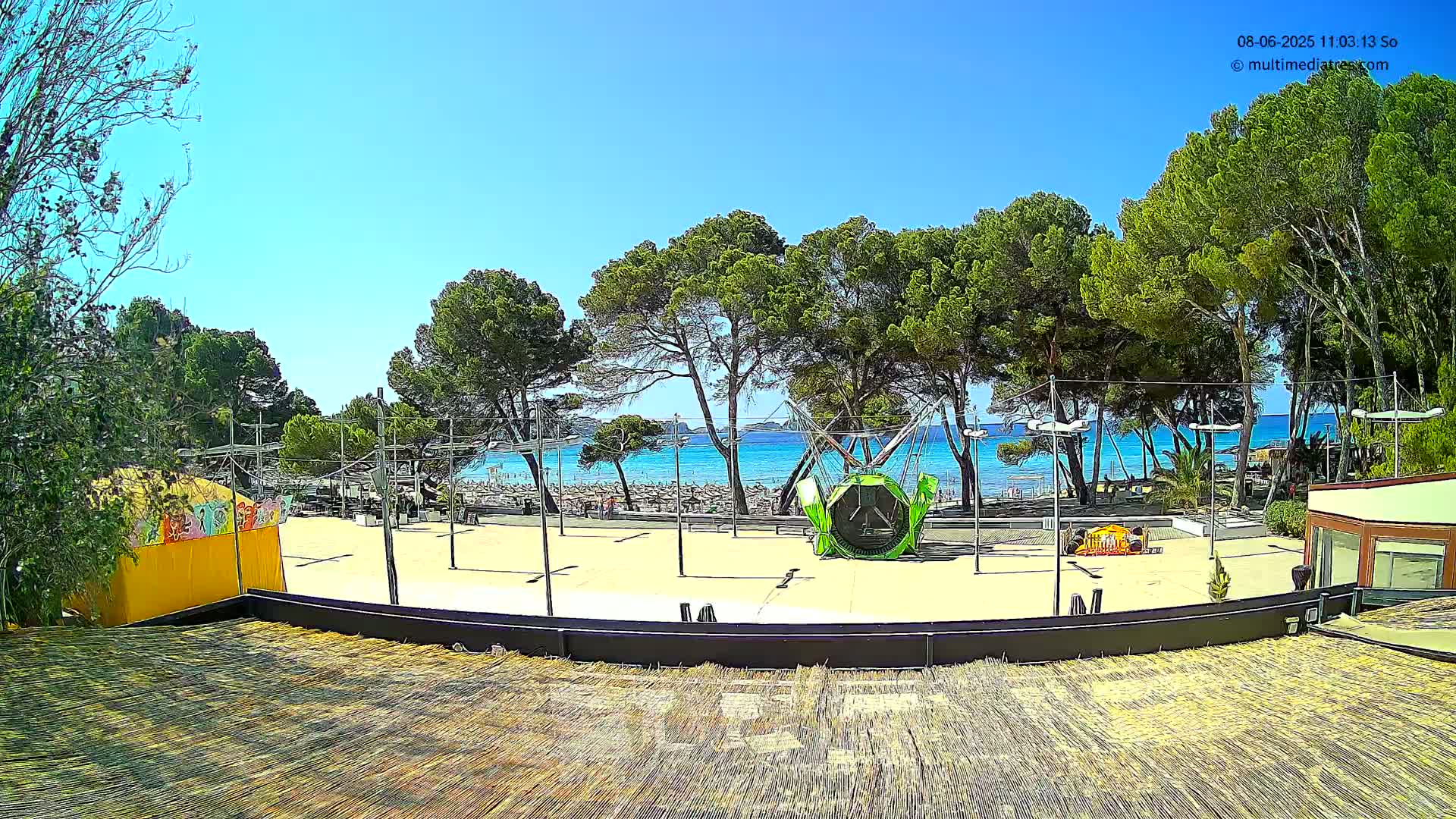 A beach with turquoise water is visible beyond a paved area with a small amusement park ride and several trees under a clear blue sky.